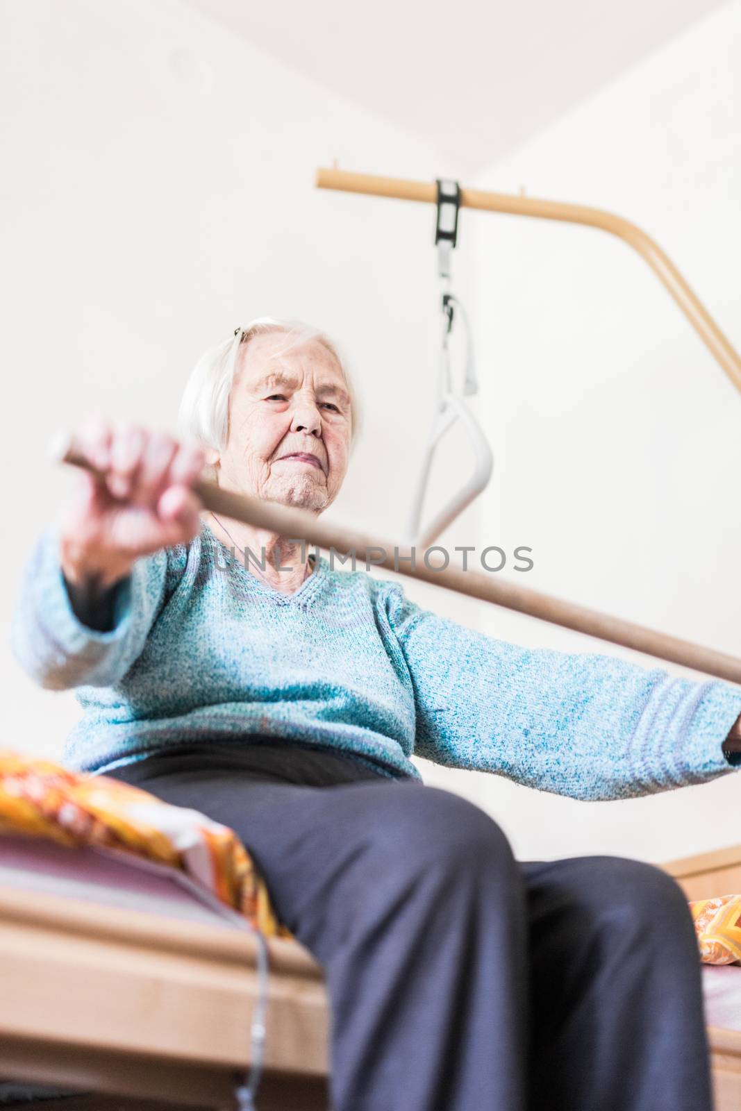 Elderly 96 years old woman exercising with a stick sitting on her bad. by kasto