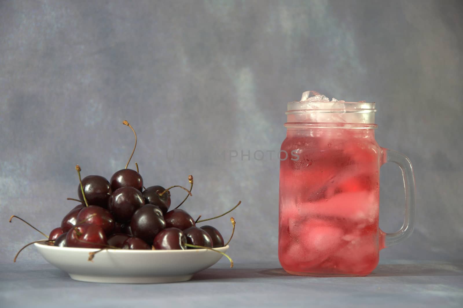 A pile of ripe cherries on a plate and a glass mug with cherry juice with ice.