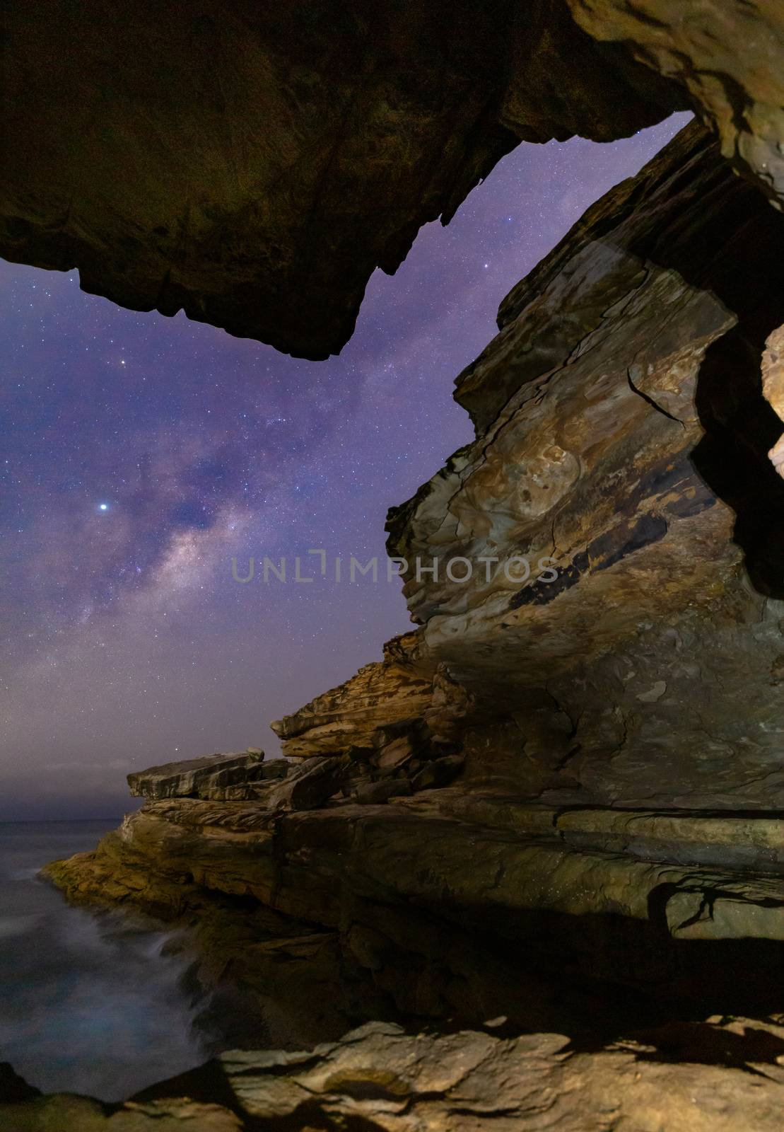 Lined up the Milky Way and galactic core rising over the Sydney sea coast down in the cliffs and aligned with the narrow gap in rock formations.  Vertical composition suitable for books, borchures or magazines