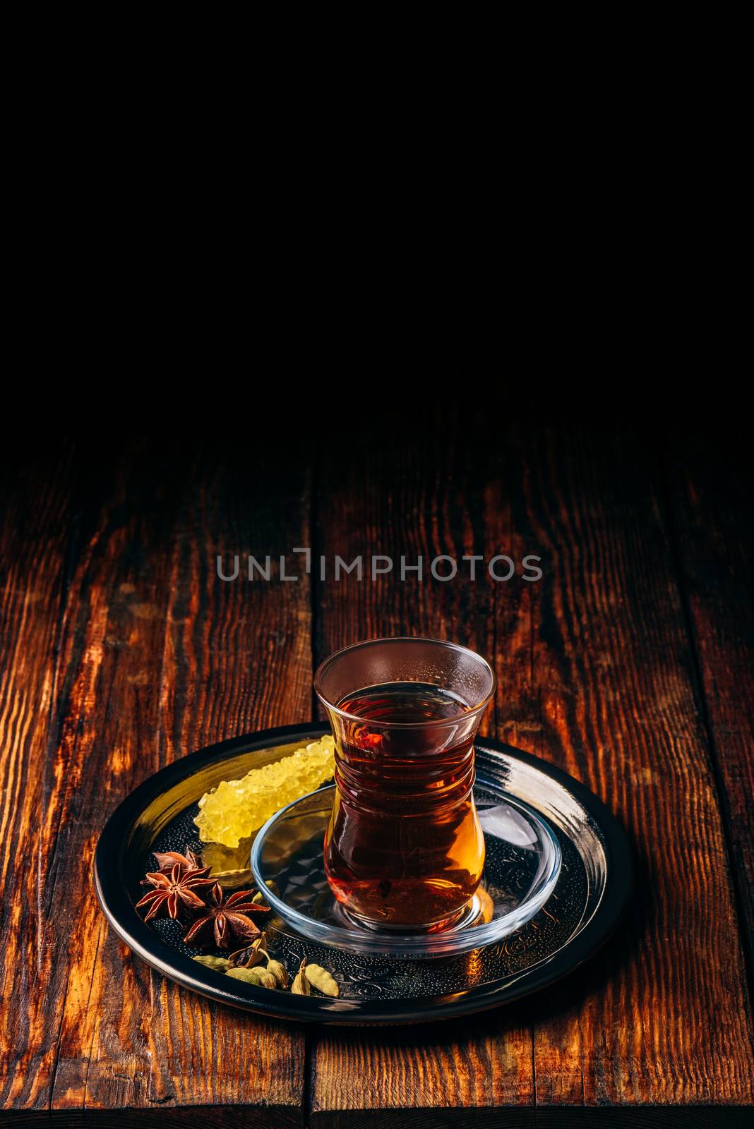 Black tea in armudu glass with star anise and green cardamom with crystal sugar on metal tray over wooden surface. Copy space