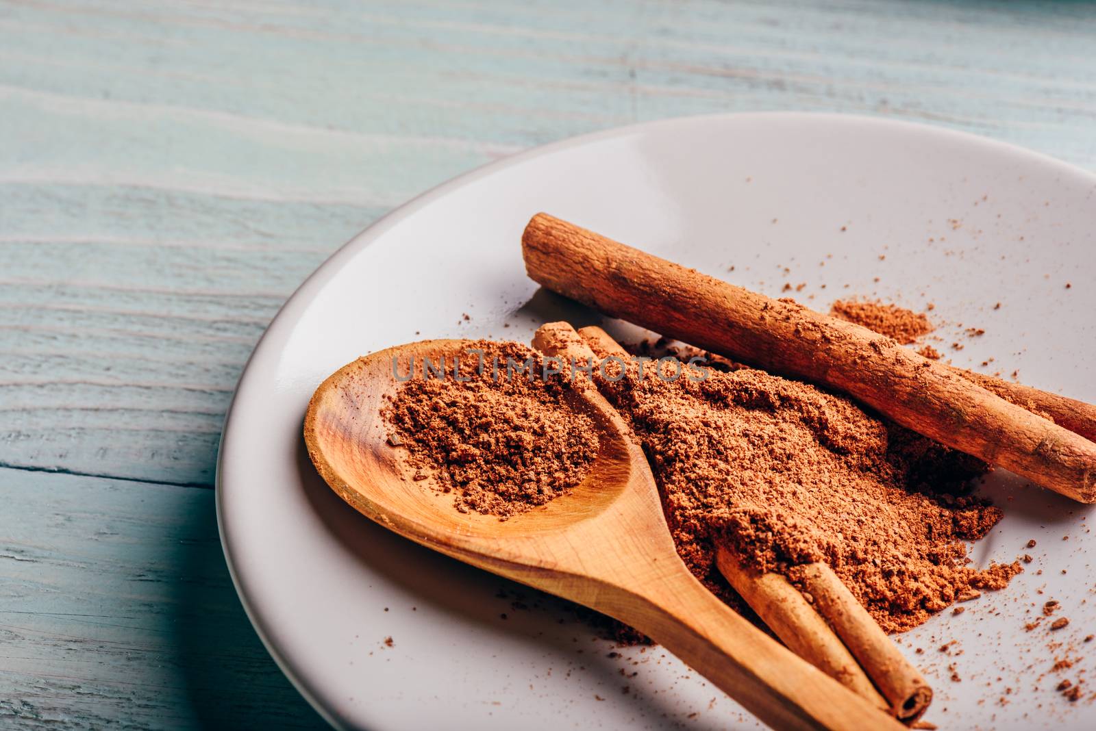 Cinnamon sticks and ground spice on white plate over light wooden background.