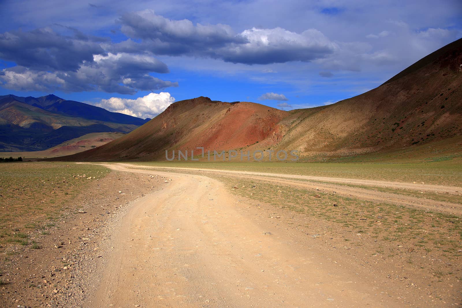 Dirt road winds at the foot of the red mountains under the thunderclouds in the sky. Altai, Siberia, Russia