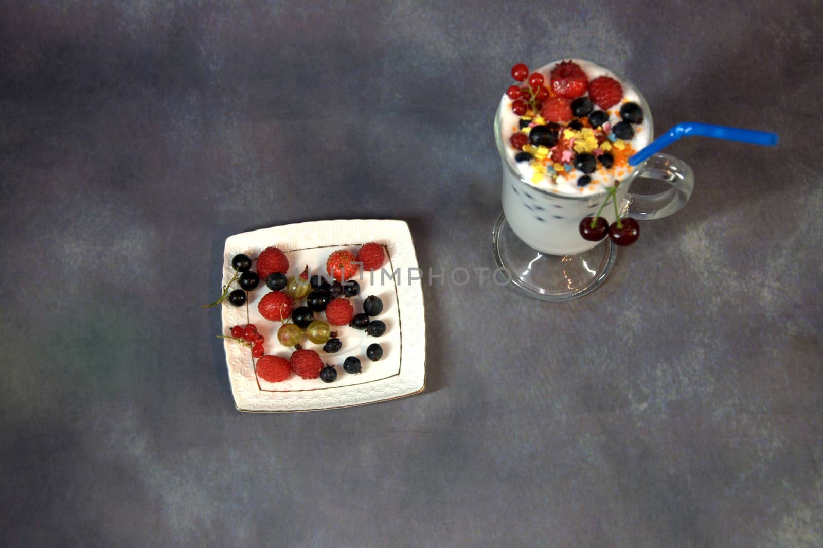 A glass with a milkshake with wild berries and a straw, stands next to a saucer filled with berries.
