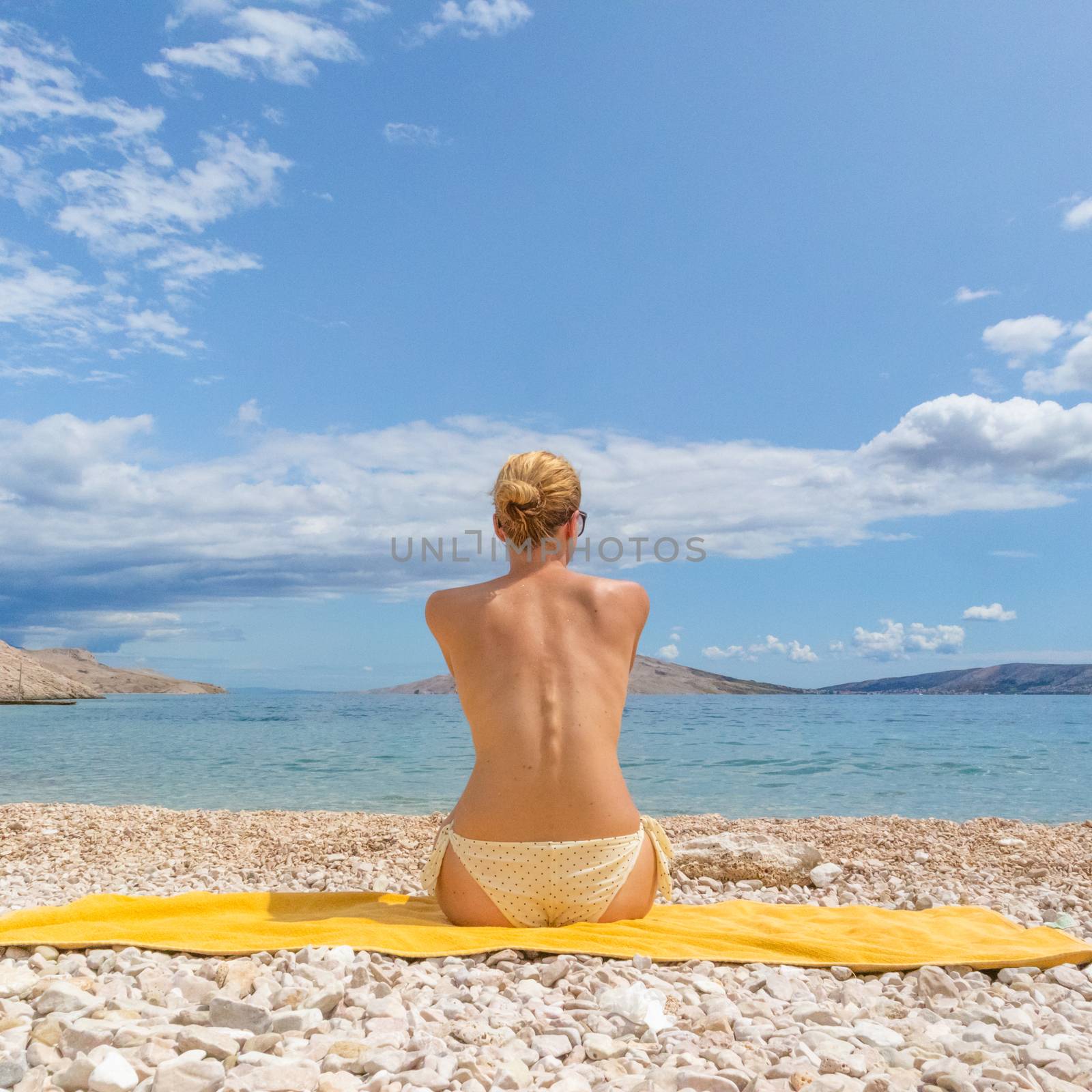 Rear view of relaxed sexy young caucasian woman sitting on yellow towel, sunbathing topless alone on romote pabble beach on Pag island, Croatia, Mediterranean. Enjoying pure nature.