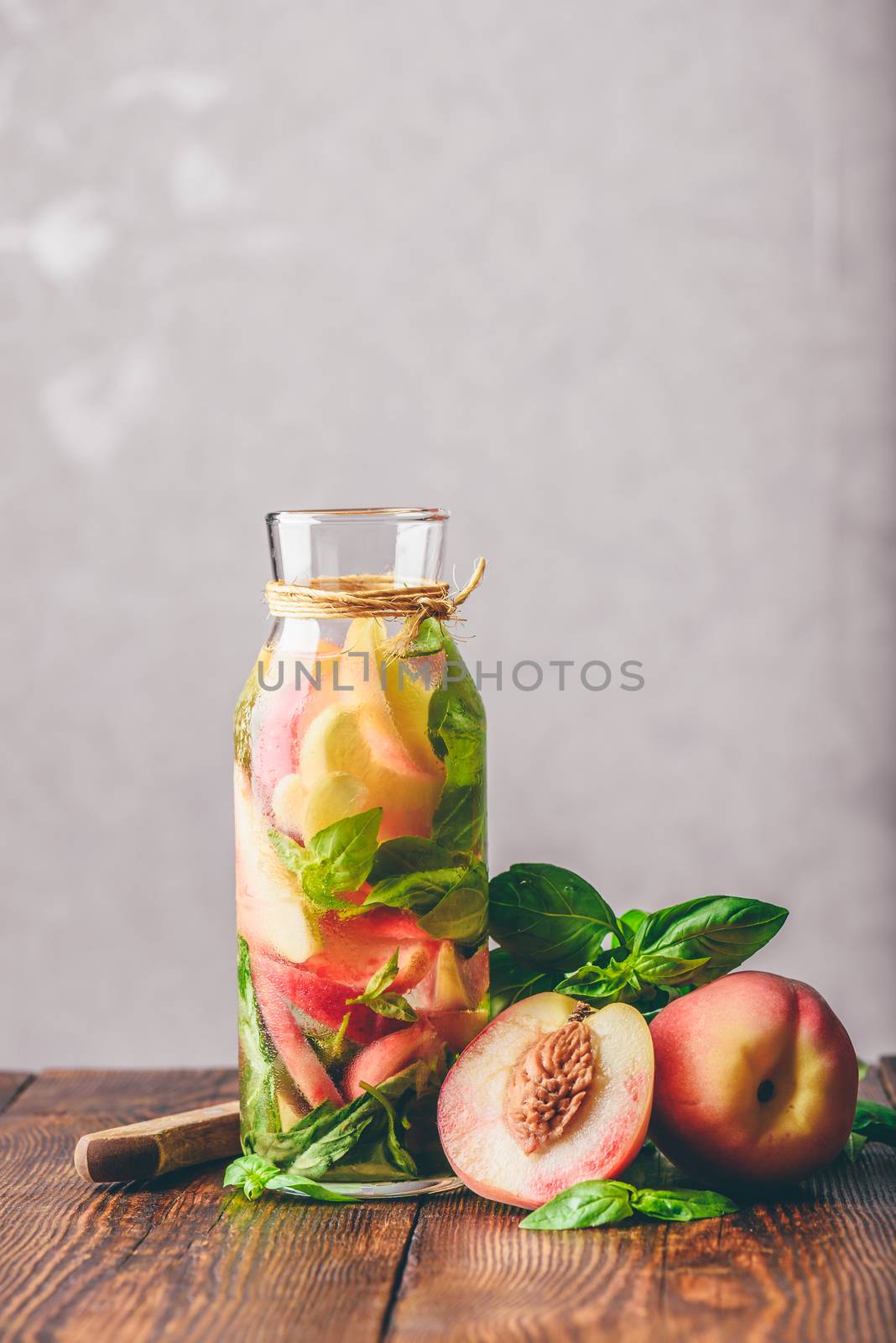 Bottle of Detox Water with Sliced Peach and Basil Leaves. Knife and Ingredients on Wooden Table. Vertical Orientation.