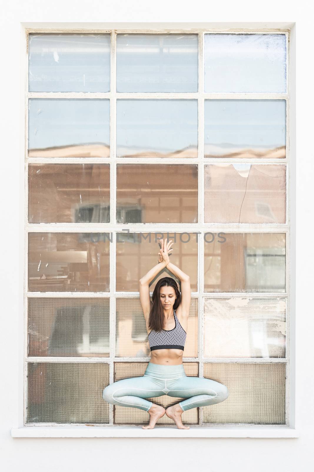 Fit sporty active girl in fashion sportswear doing yoga fitness exercise in front of big industrial window frame. colorful reflections in window glass. Outdoor sports, urban style yoga.