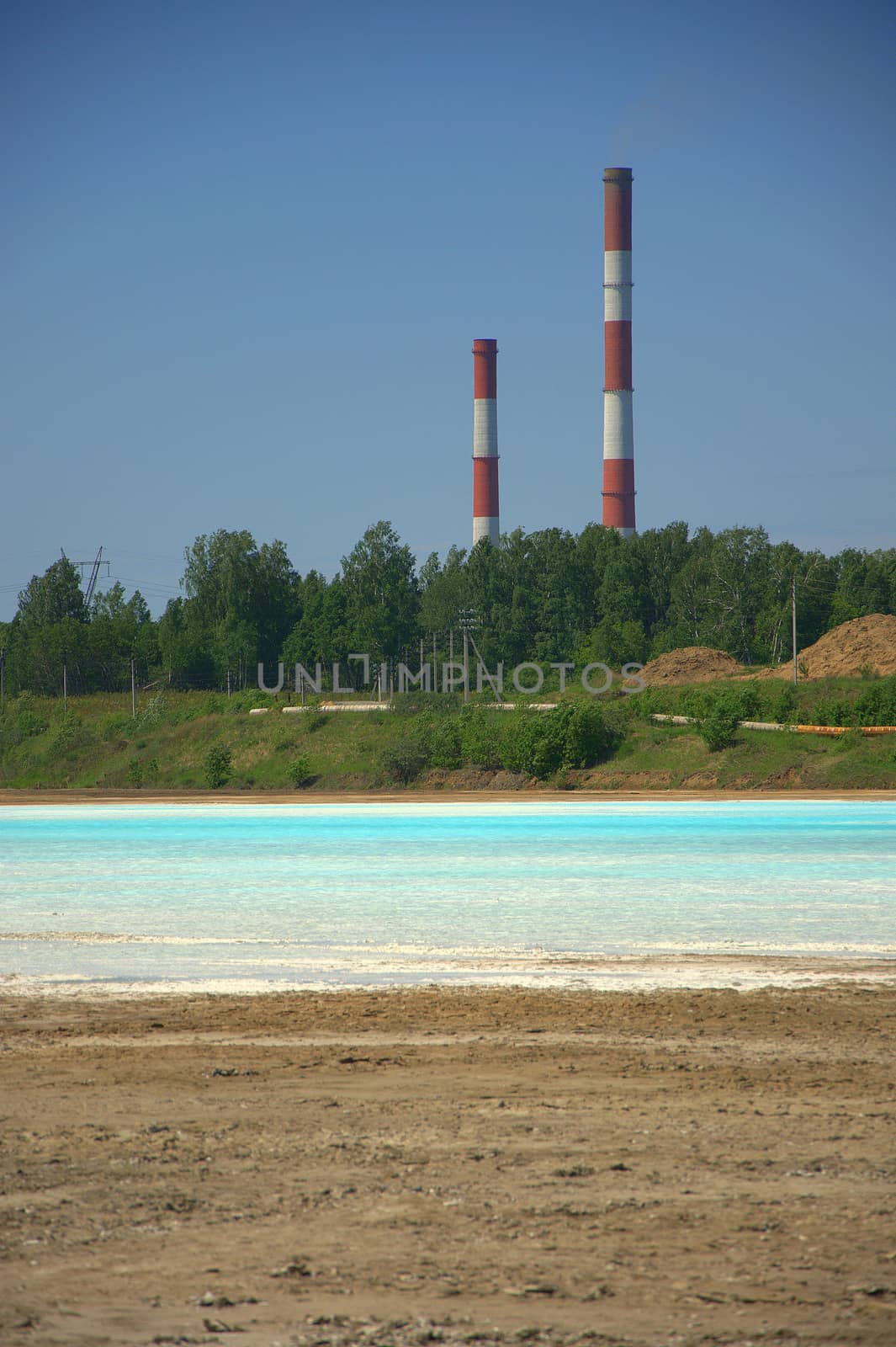 A sandy beach by a blue lake on the banks of a coniferous forest and 2 pipes of an old factory in the background.