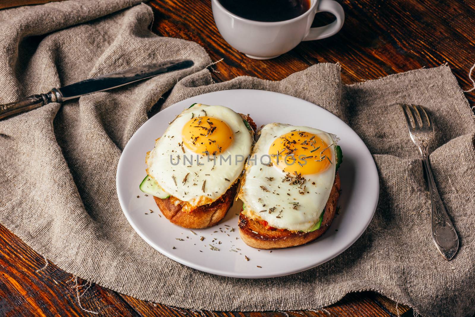 Italian toasts with vegetables and fried eggs on white plate and cup of coffee over grey rough cloth.