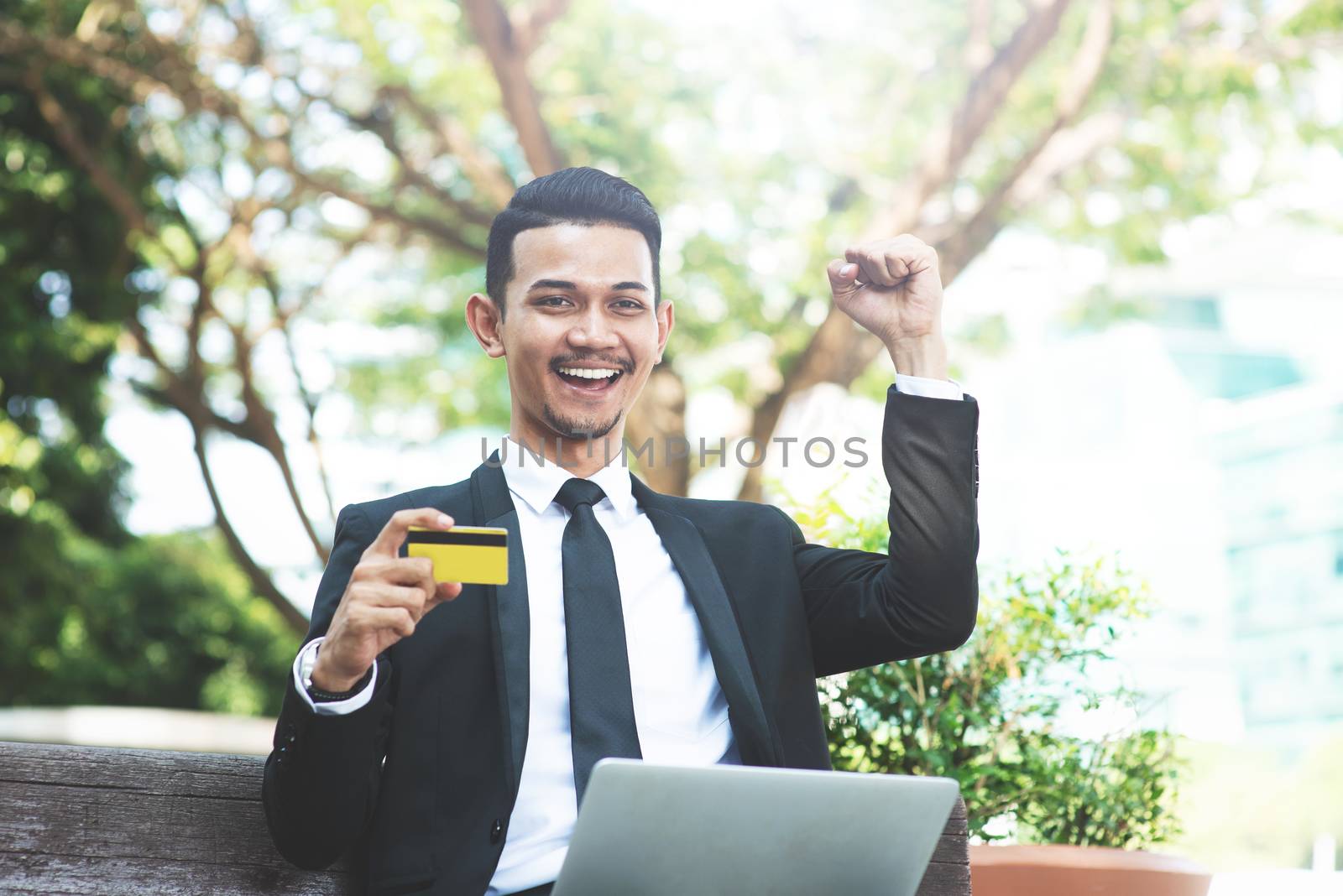 Happy Asian business man cheering, hand holding credit card sitting with laptop outdoor.