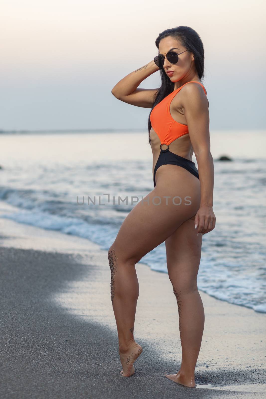 Fitness girl posing 
in the shore of the beach with a beautiful black and orange bikini and sunglasses