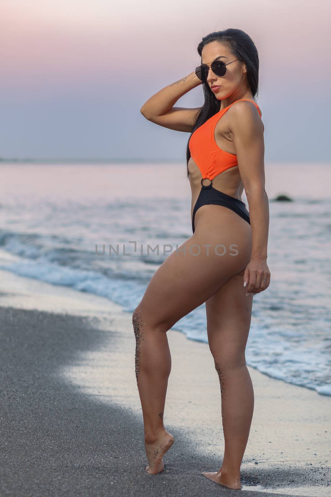 Fitness girl posing 
in the shore of the beach with a beautiful black and orange bikini and sunglasses