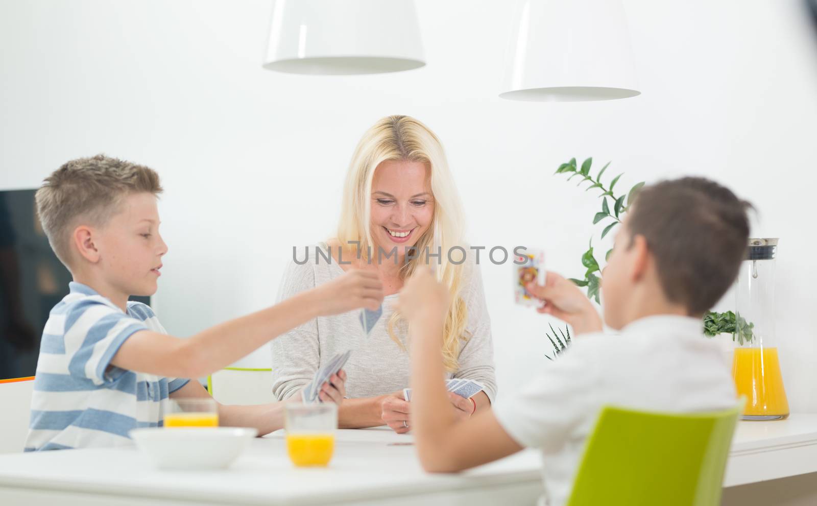 Mother and two kids playing card game at dining table at bright modern home. Spending quality leisure time with children and family concept. Cards are generic and debranded.