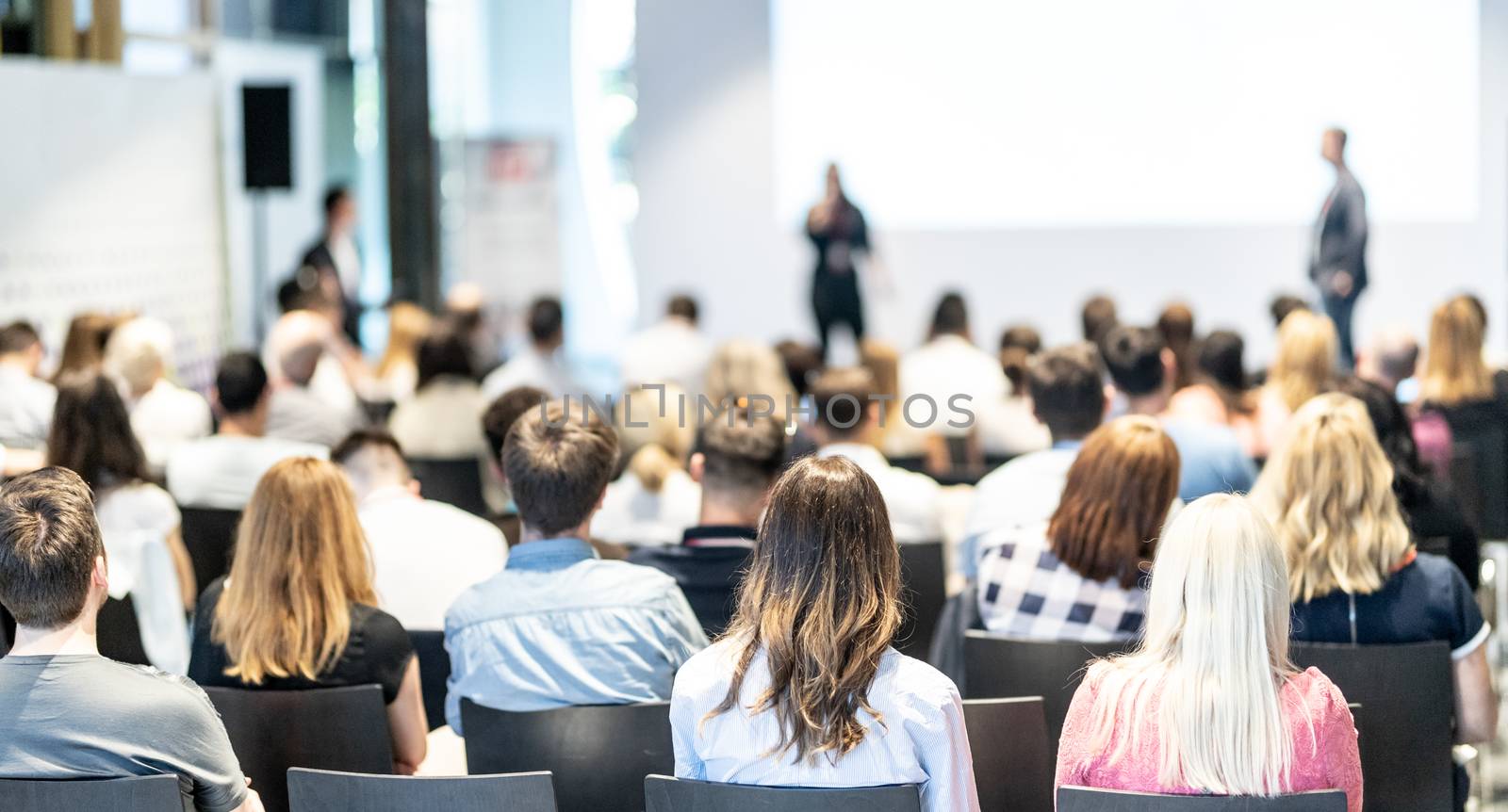 Business and entrepreneurship symposium. Speaker giving a talk at business meeting. Audience in conference hall. Rear view of unrecognized participant in audience.