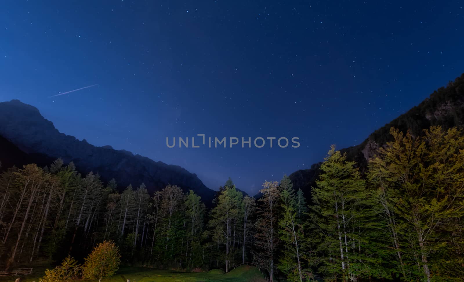 Alpine valley at night, landscape lit by full moon, stars visible, forest in front, trails of airplane above mountains