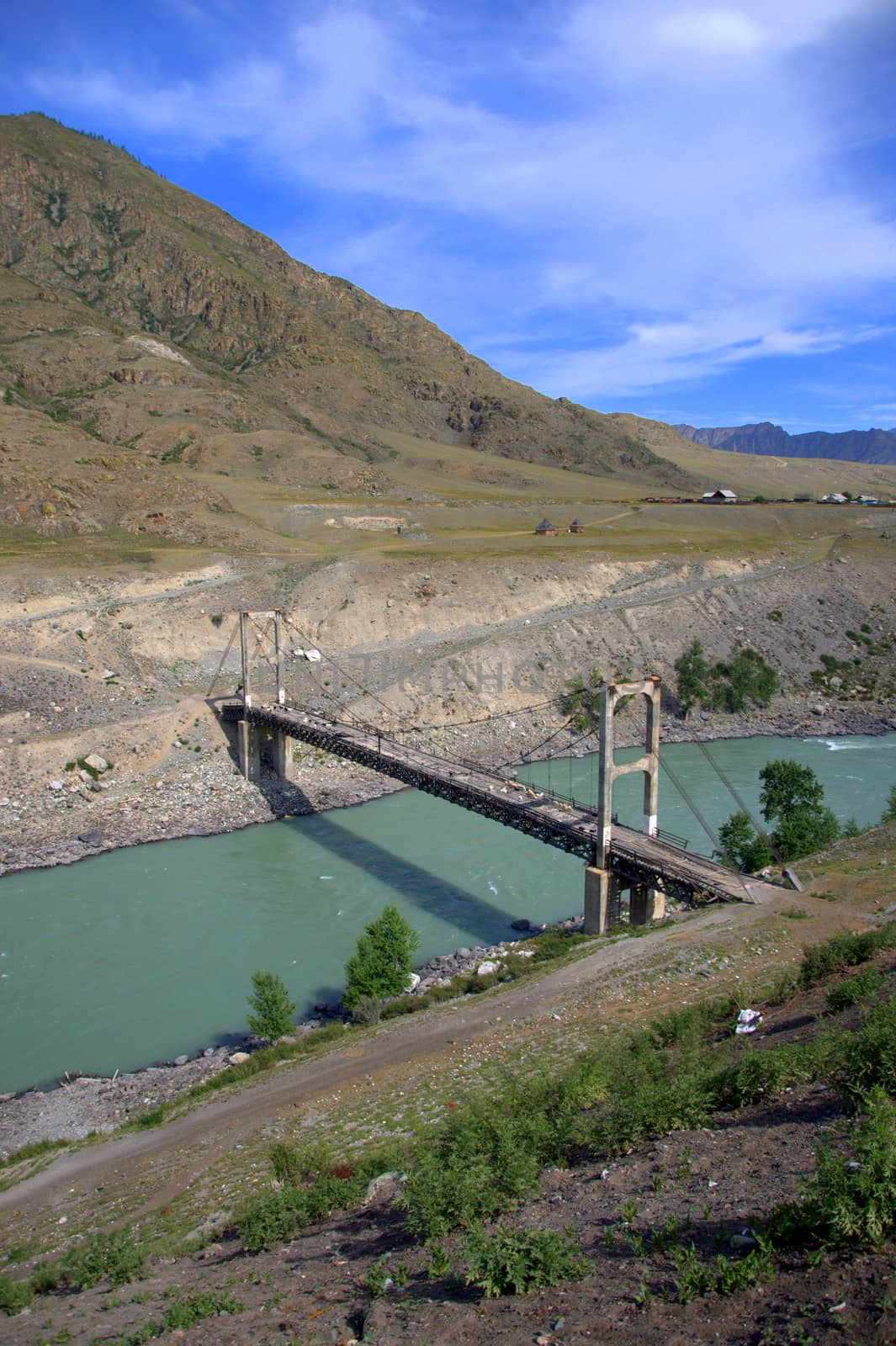 Old cable-stayed bridge over a turquoise river against the backdrop of mountain ranges. Altai, Siberia, Russia.