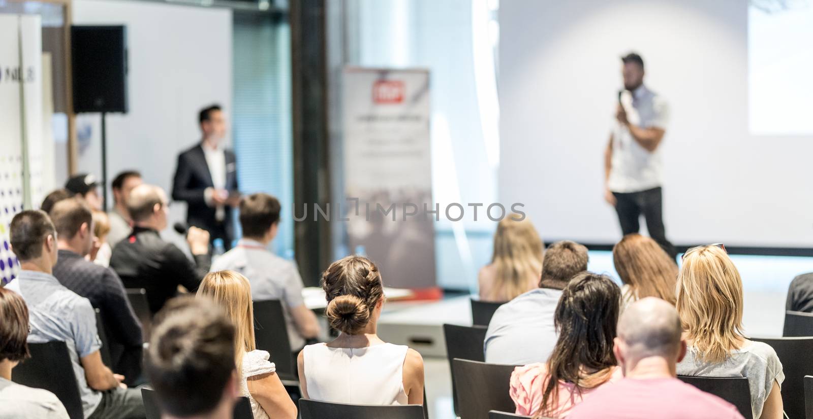 Business and entrepreneurship symposium. Speaker giving a talk at business meeting. Audience in conference hall. Rear view of unrecognized participant in audience.