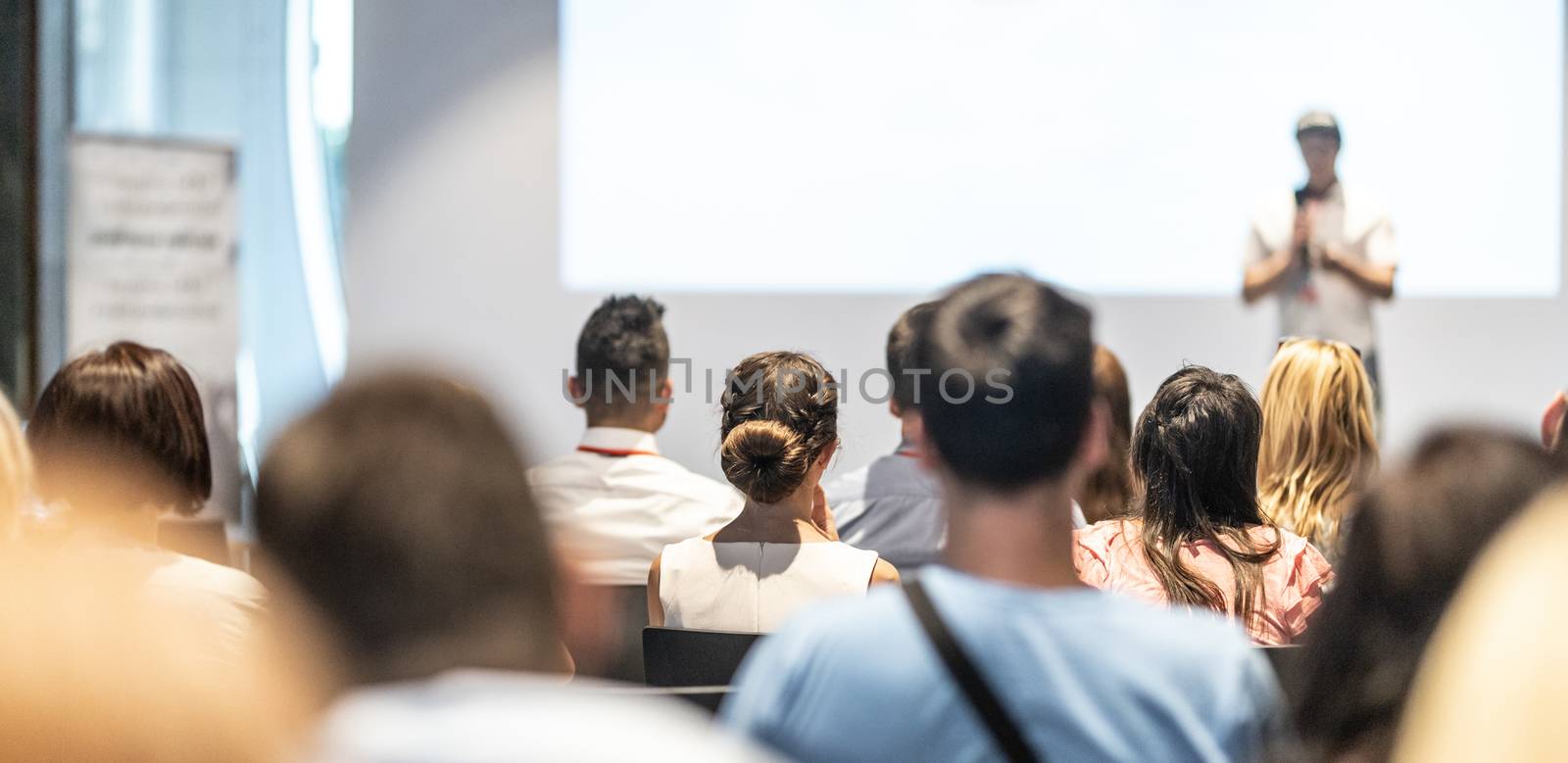 Business and entrepreneurship symposium. Speaker giving a talk at business meeting. Audience in conference hall. Rear view of unrecognized participant in audience.