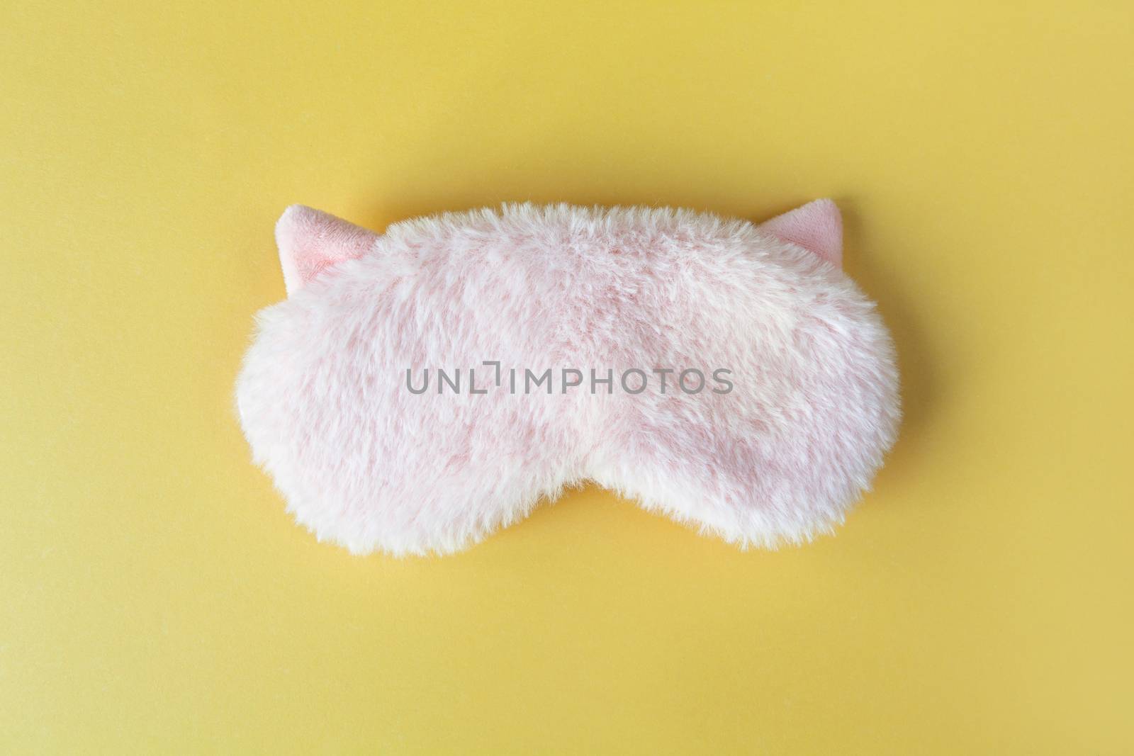 Pastel pink fluffy fur sleep mask with small ears on pastel yellow paper background. Top view, flat lay. Concept of vivid dreams. Accessories for girls and young women.