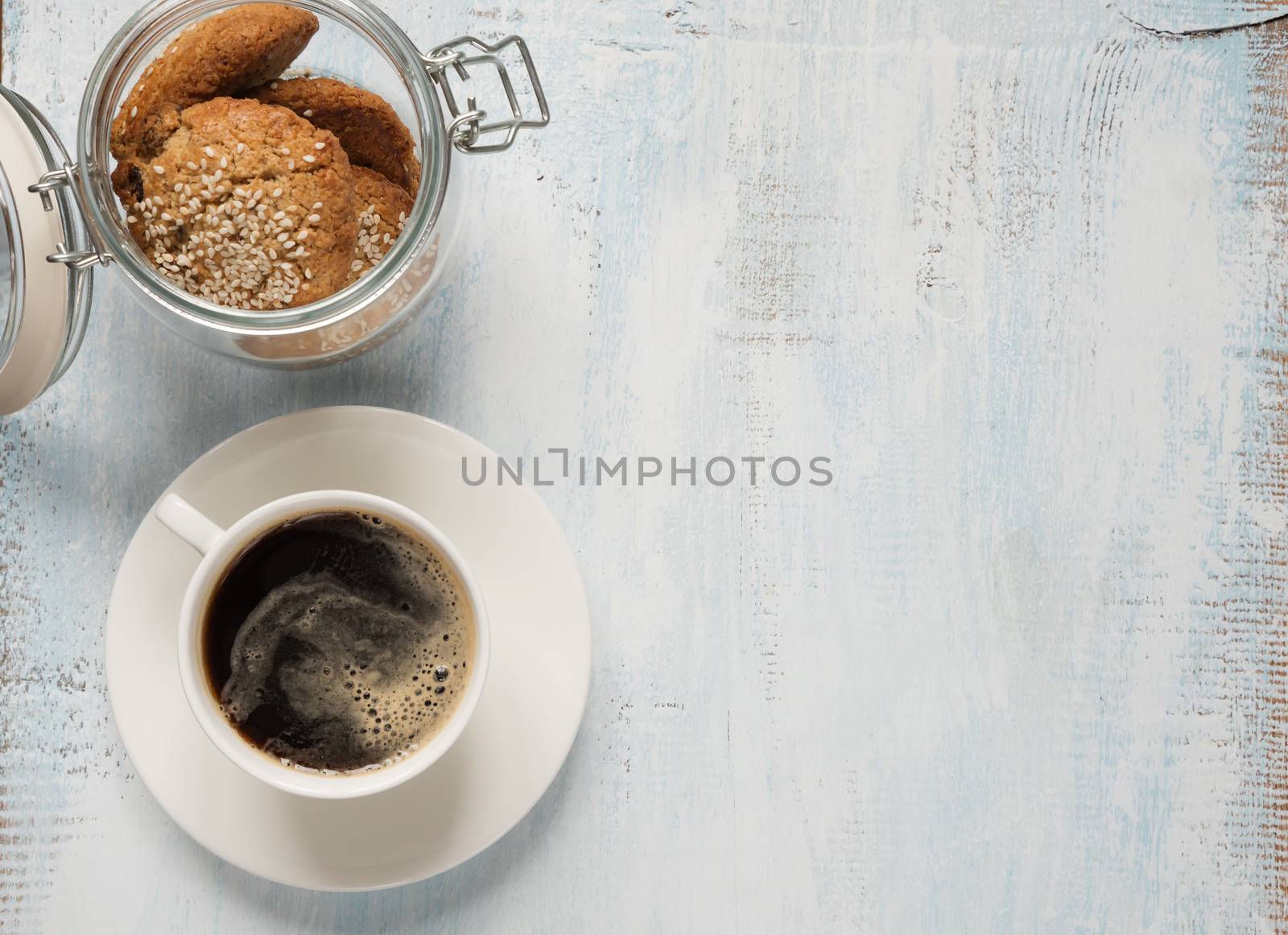 Black coffee with crema in white porcelain cup and oatmeal cookie in a glass jar on a light wooden background with space for text, top view