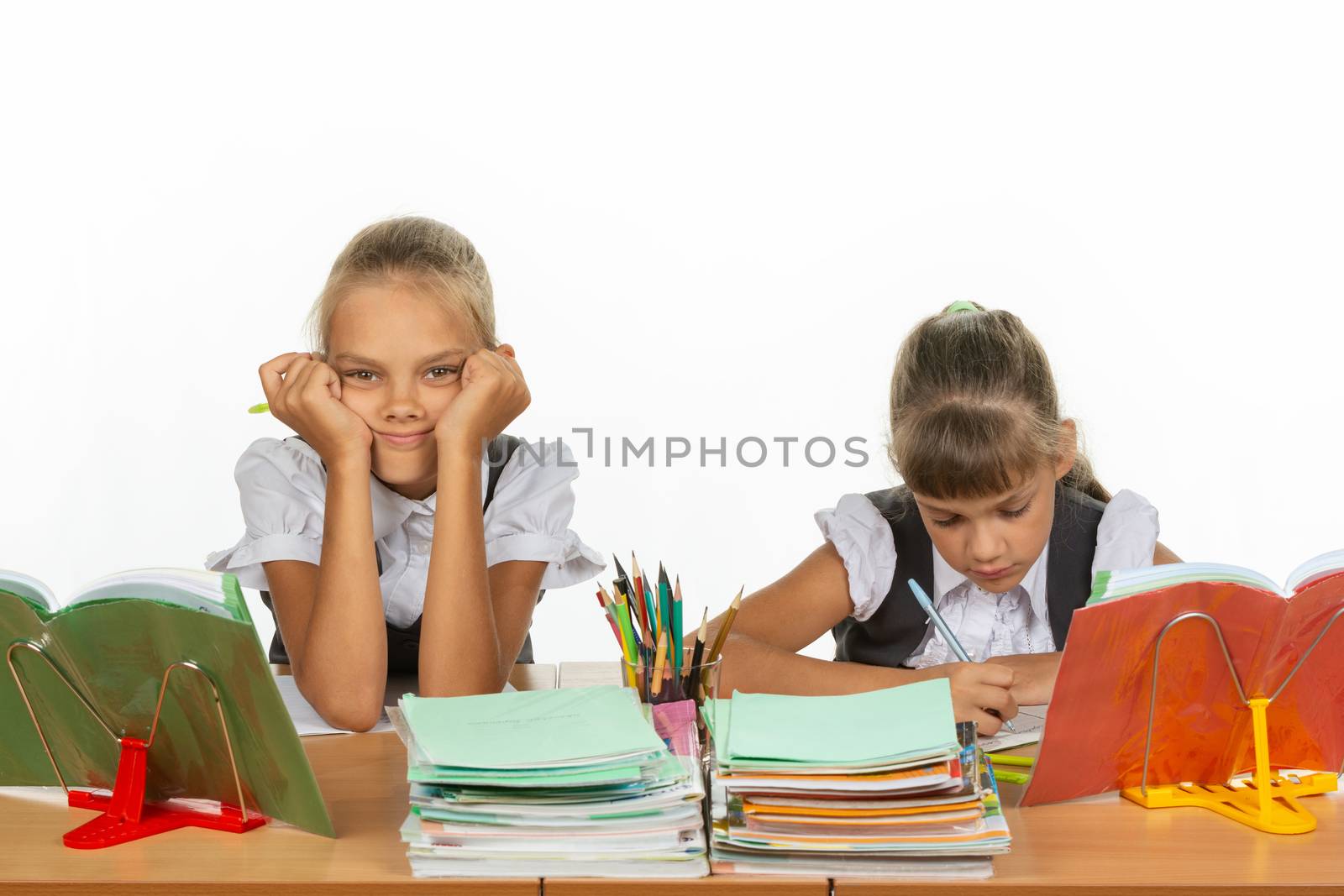 Two schoolgirls at a desk, one leaning on her hands and looking into the frame, the other writes