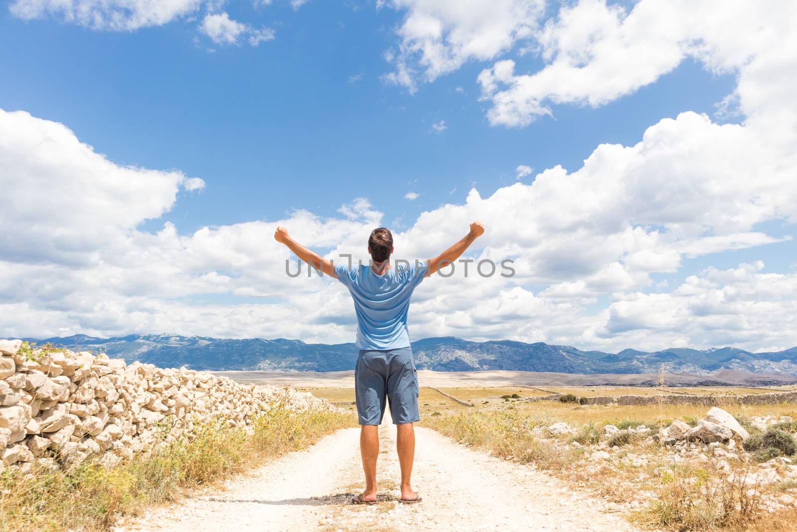 Rear view of casual sporty man standing on a dirt country road rising hands up to the clouds on a blue summer sky. Freedom and travel adventure concept. Copy space.