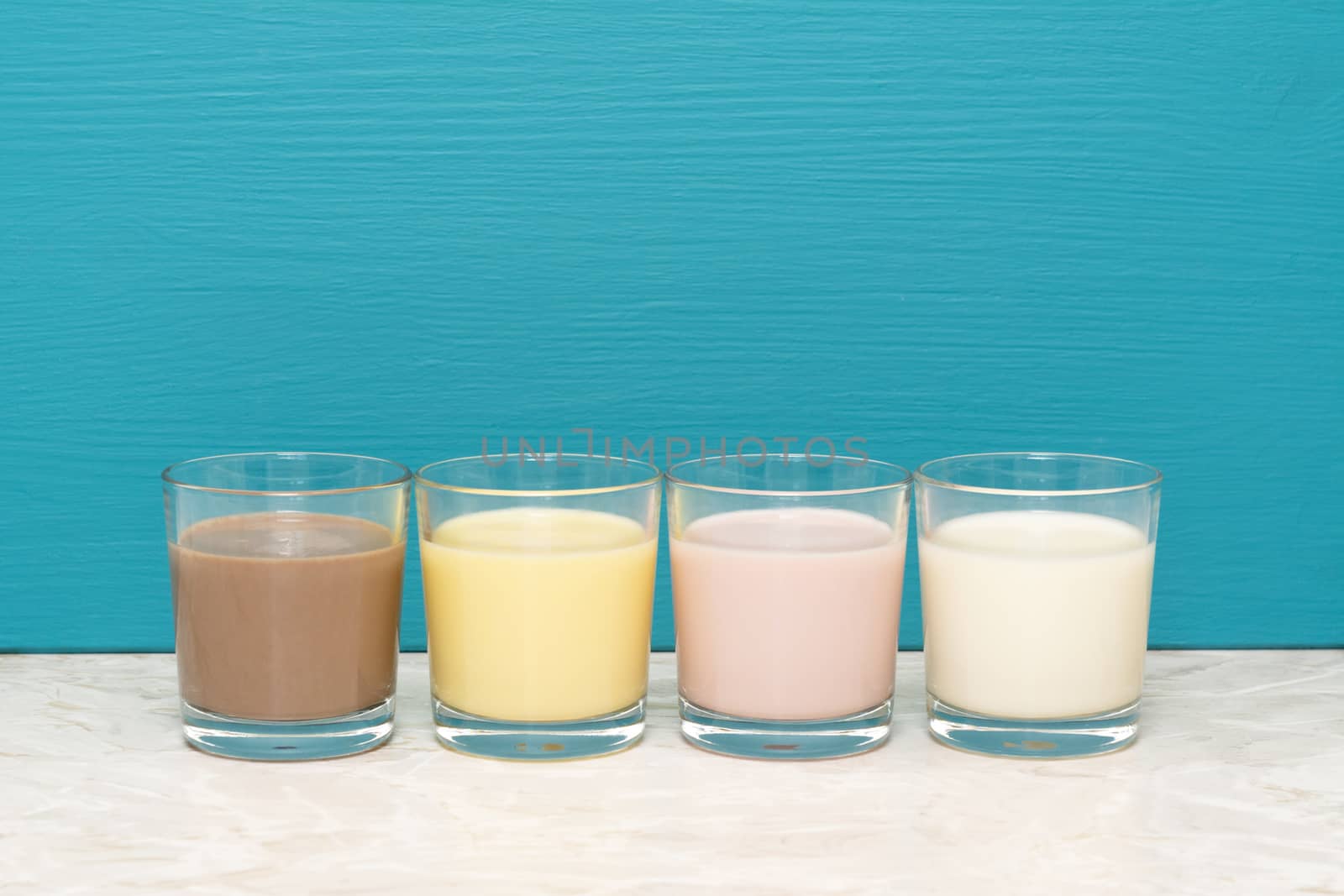 Chocolate, banana and strawberry milkshakes and fresh milk in glass tumblers against a teal background