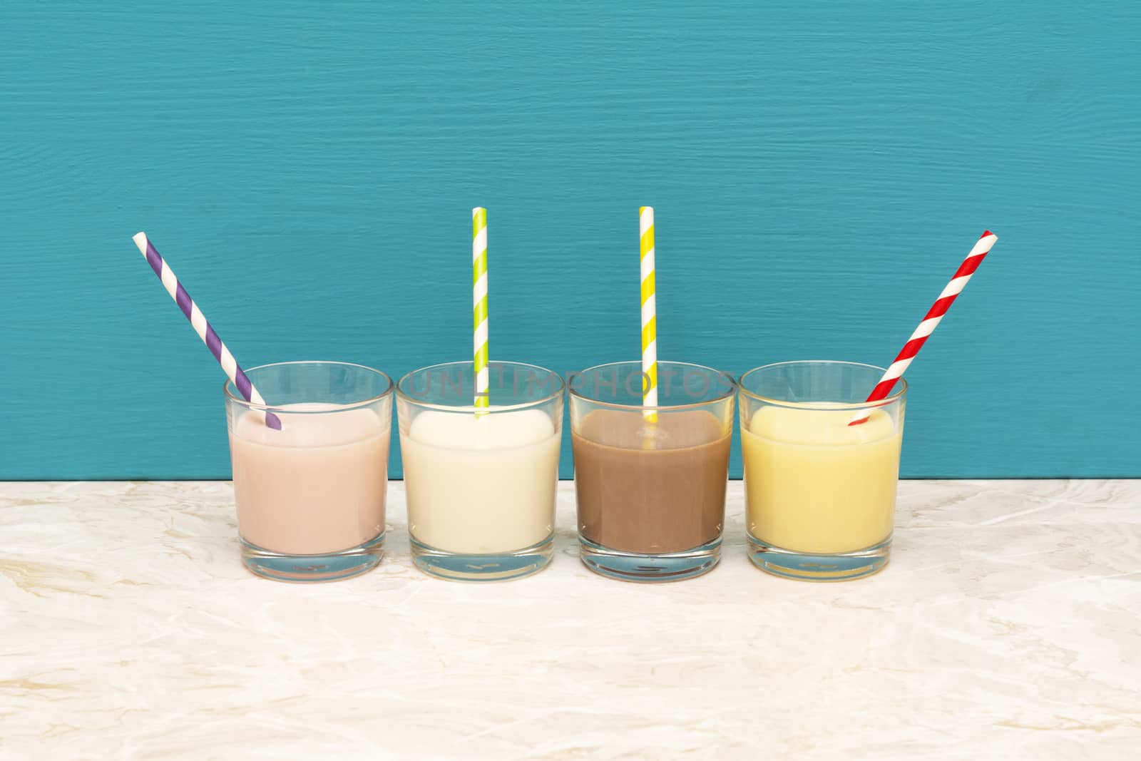 Flavoured milkshakes - strawberry, chocolate and banana - and fresh creamy milk with retro paper straws in glass tumblers with a teal background