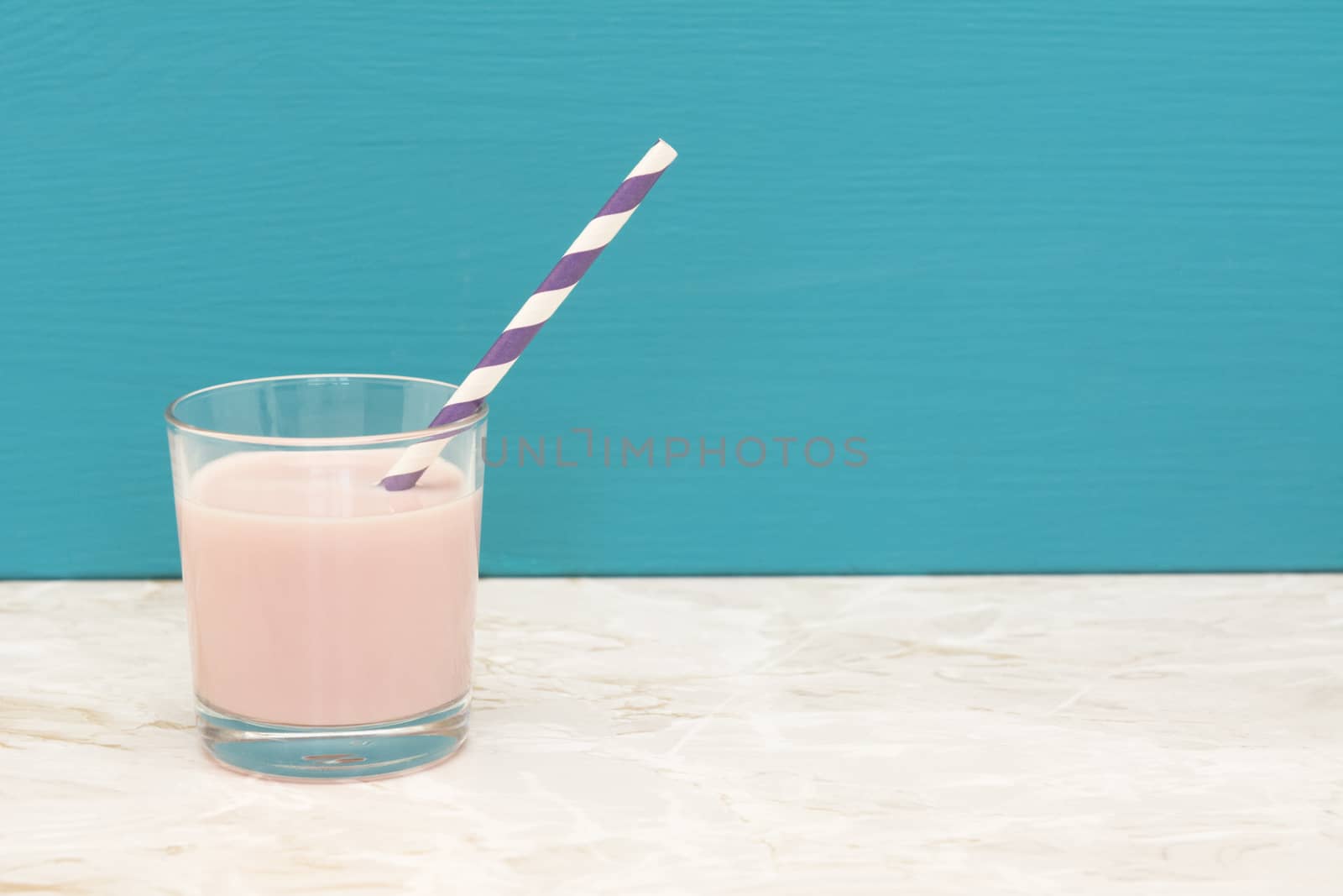 Delicious strawberry milkshake with a retro paper straw in a glass tumbler with a teal background and copy space