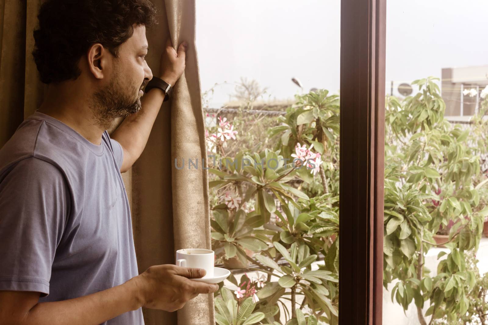 Man holding a tasty cup of sweet smelling morning coffee, relaxing and looking through the window. Morning sun light coming from outside. Happy weekend concept. Copy space room for text on left.