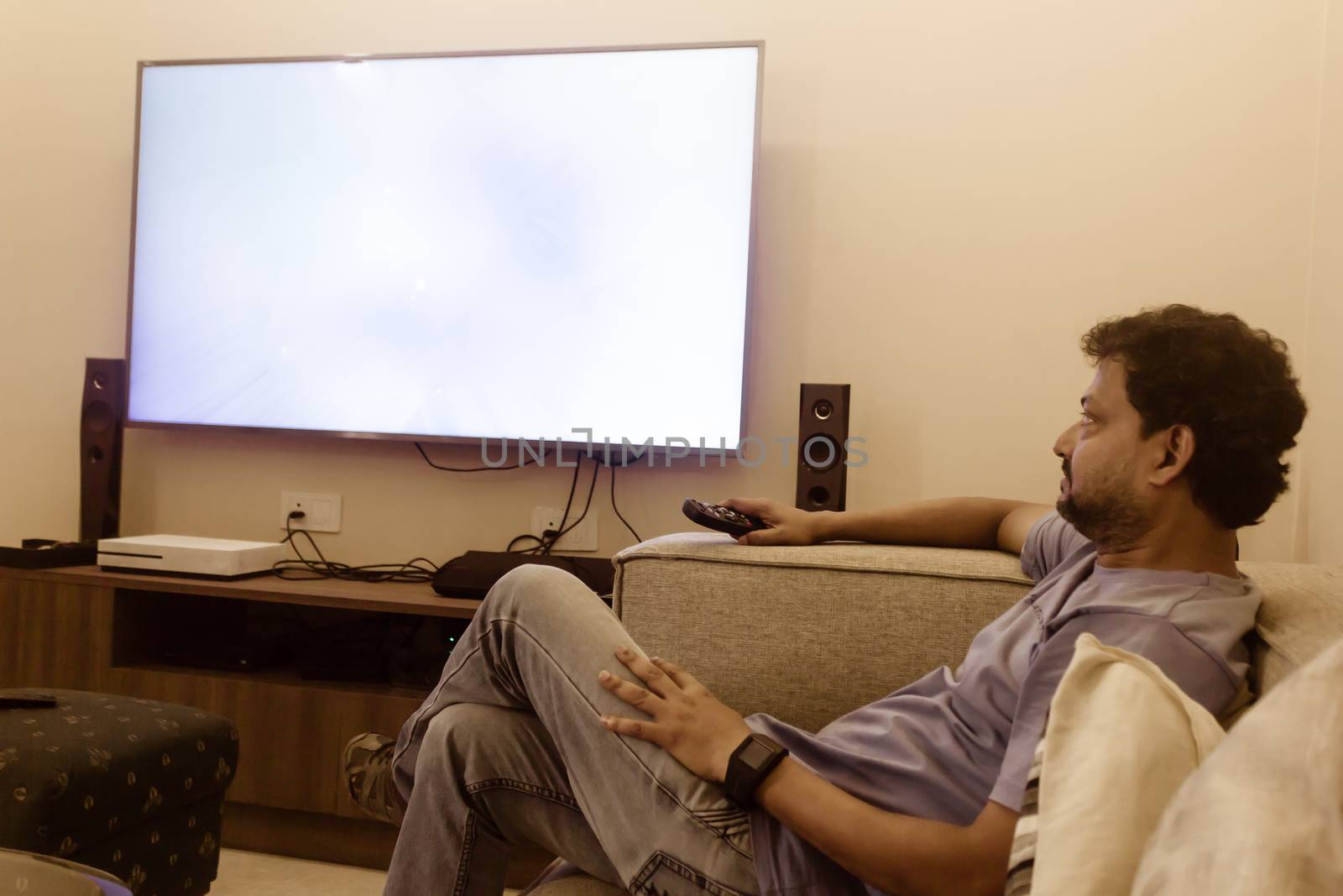 Man watching tv at home in living room. Asian man in blue T shirt sitting down on a couch watching television on sofa. side view. Leisure, technology, mass media and people concept by sudiptabhowmick