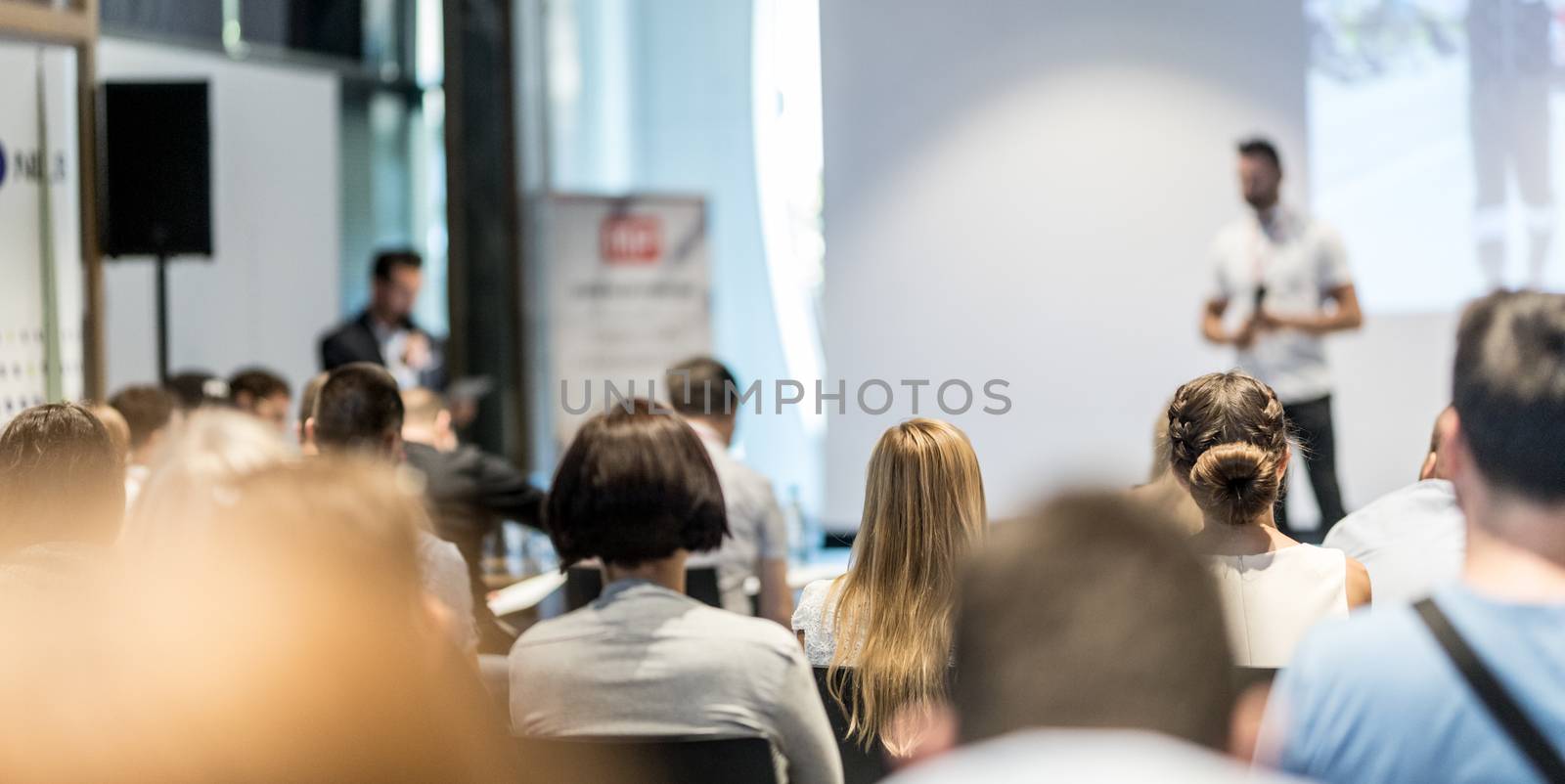 Business and entrepreneurship symposium. Speaker giving a talk at business meeting. Audience in conference hall. Rear view of unrecognized participant in audience.