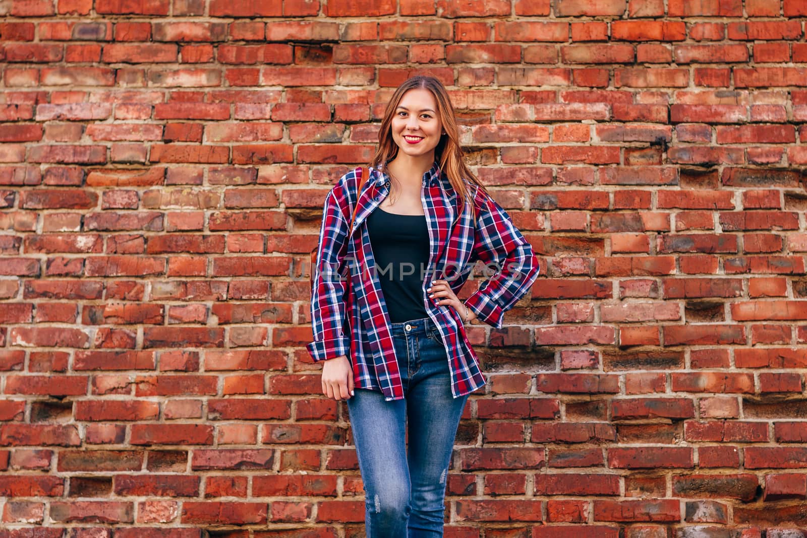 Portrait of young woman in checkered shirt and blue jeans standing against red brick wall