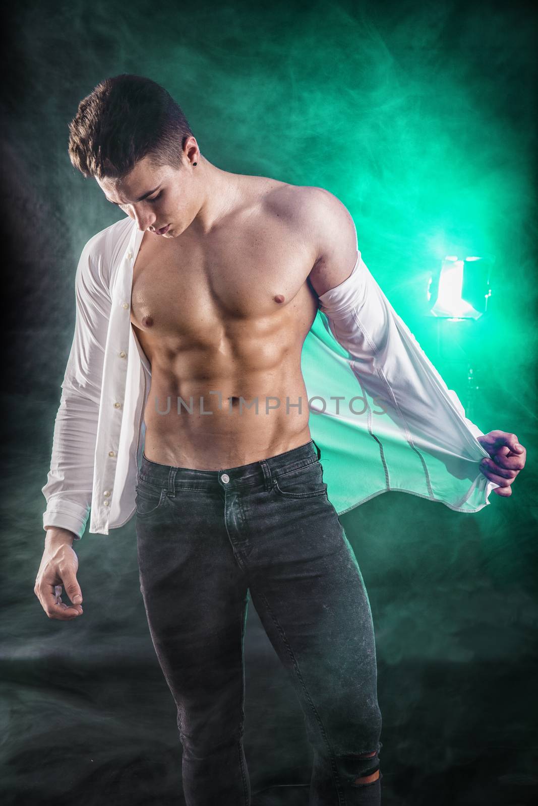 Handsome young muscular man shirtless wearing jeans, taking off leather jacket on naked muscle torso, on dark background in studio shot