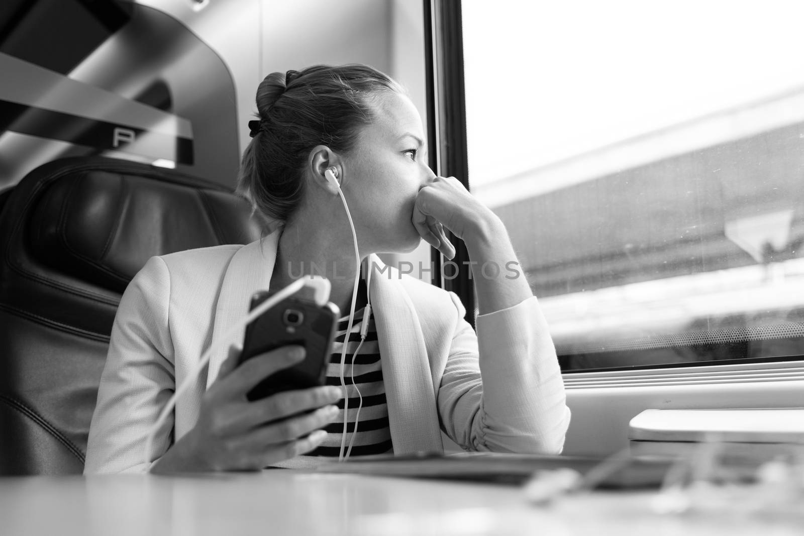 Thoughtful businesswoman looking trough the window, listening to podcast on cellphone using headphone set while traveling by train in business class seat. Black and white image.