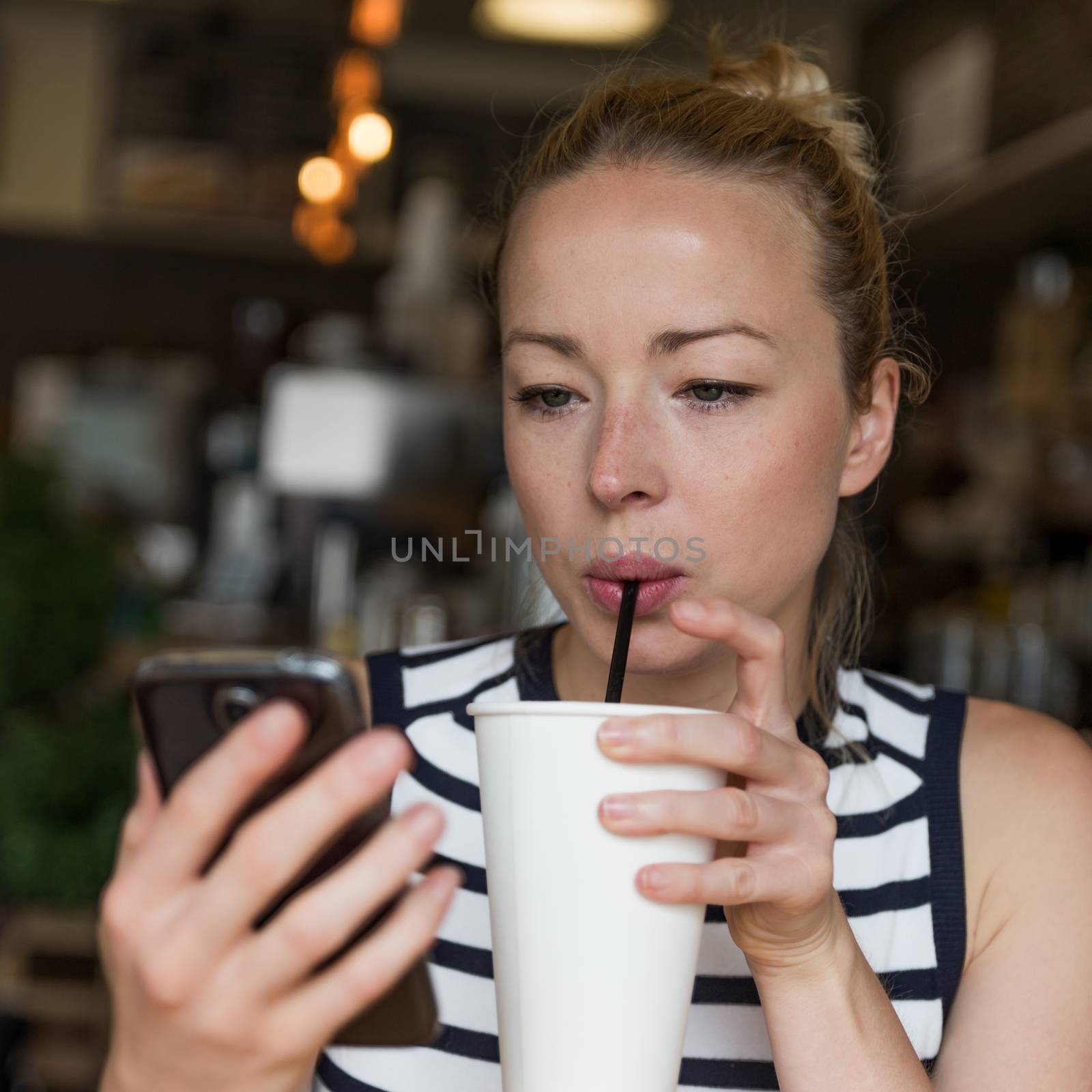 Thoughtful woman reading news on mobile phone during rest in coffee shop. Happy Caucasian female watching her photo on cell telephone while sipping coffee in cafe during free time.