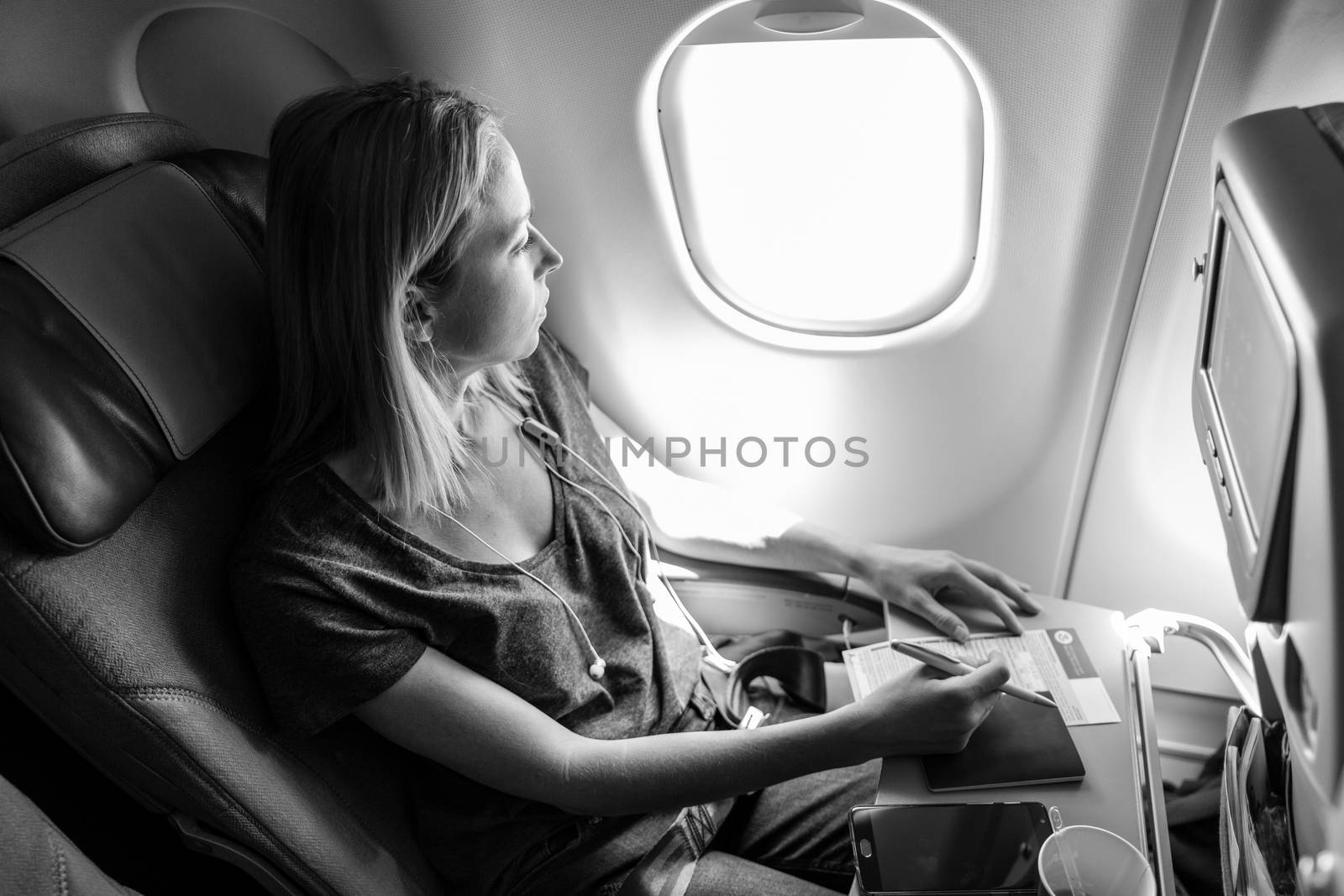 Relaxed casual woman flying on commercial passengers airplane, filling in immigration form, drinking coffee, listening to music, looking through the window. Black and white image.