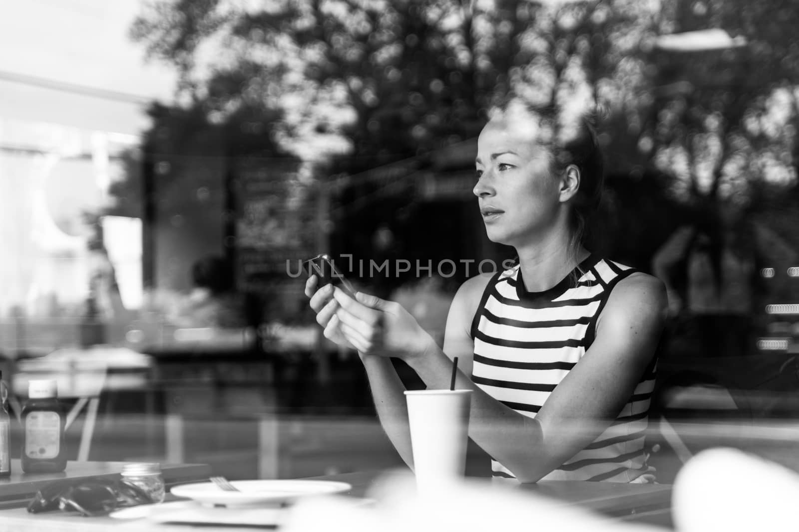 Thoughtful caucasian woman holding mobile phone while looking through the coffee shop window during coffee break. Street reflections in the window glass. Black and white image.