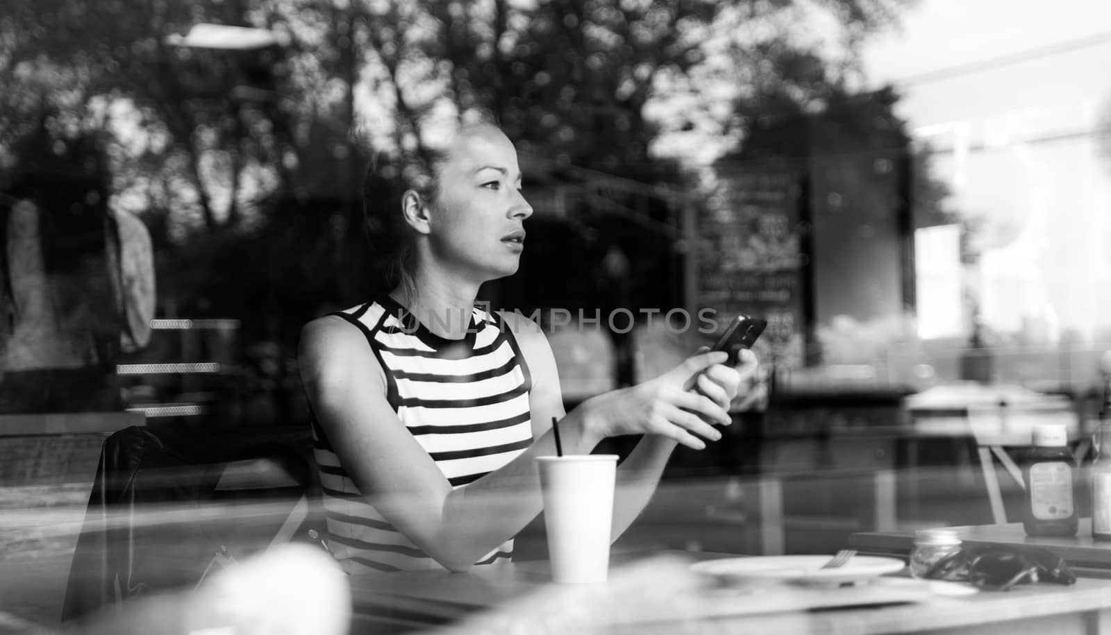 Thoughtful caucasian woman holding mobile phone while looking through the coffee shop window during coffee break. Street reflections in the window glass. Black and white image.