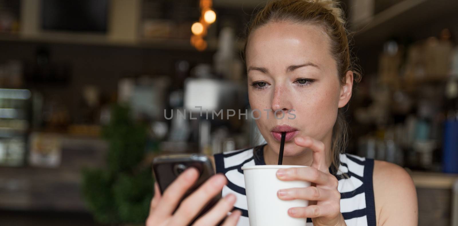 Thoughtful woman reading news on mobile phone during rest in coffee shop. Happy Caucasian female watching her photo on cell telephone while sipping coffee in cafe during free time.