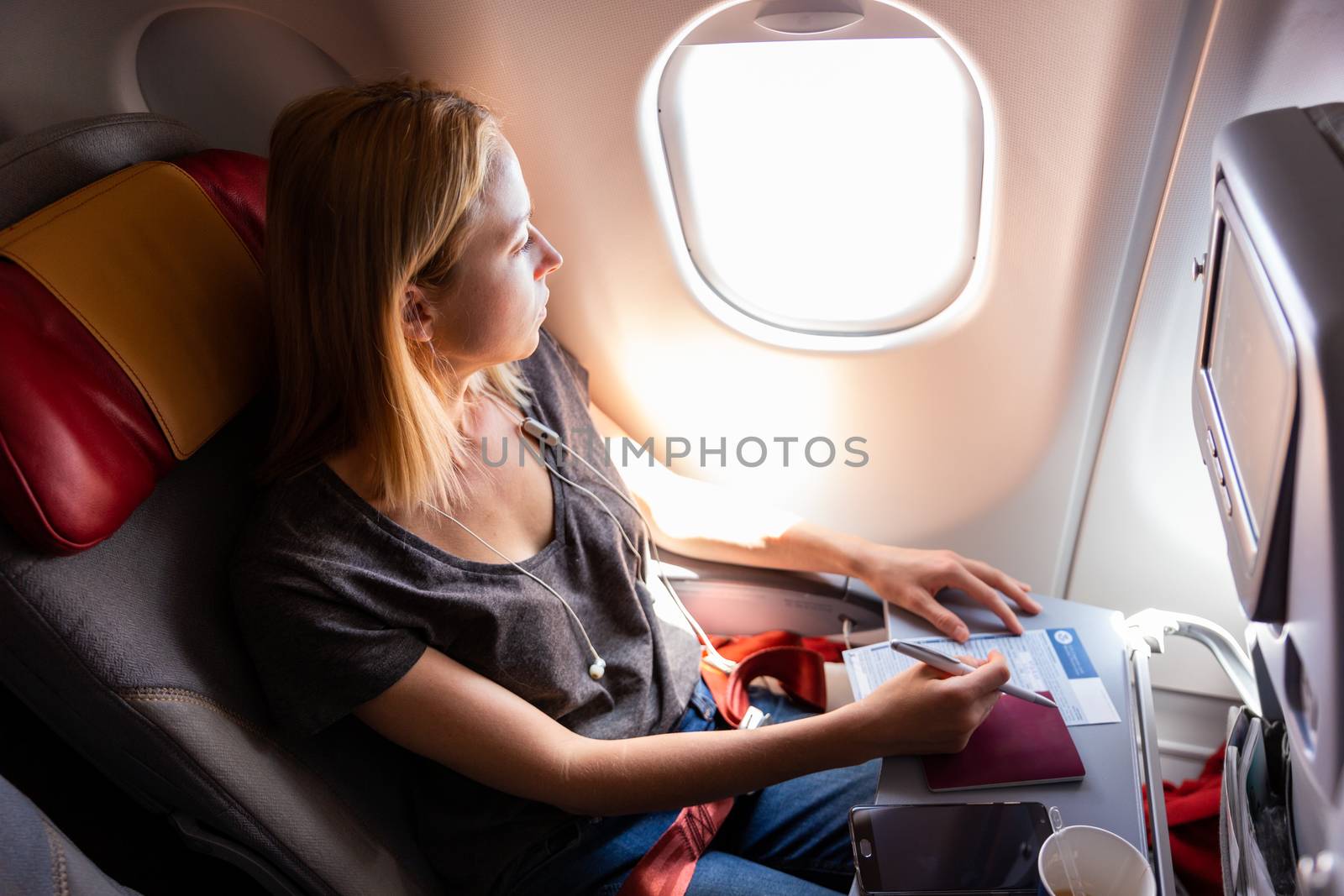 Relaxed casual woman flying on commercial passengers airplane, filling in immigration form, drinking coffee, listening to music, looking through the window.