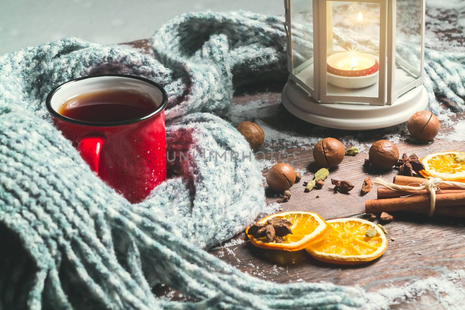 Lantern with a burning candle, spices and a red mug with hot coffee on a snowy wooden table.