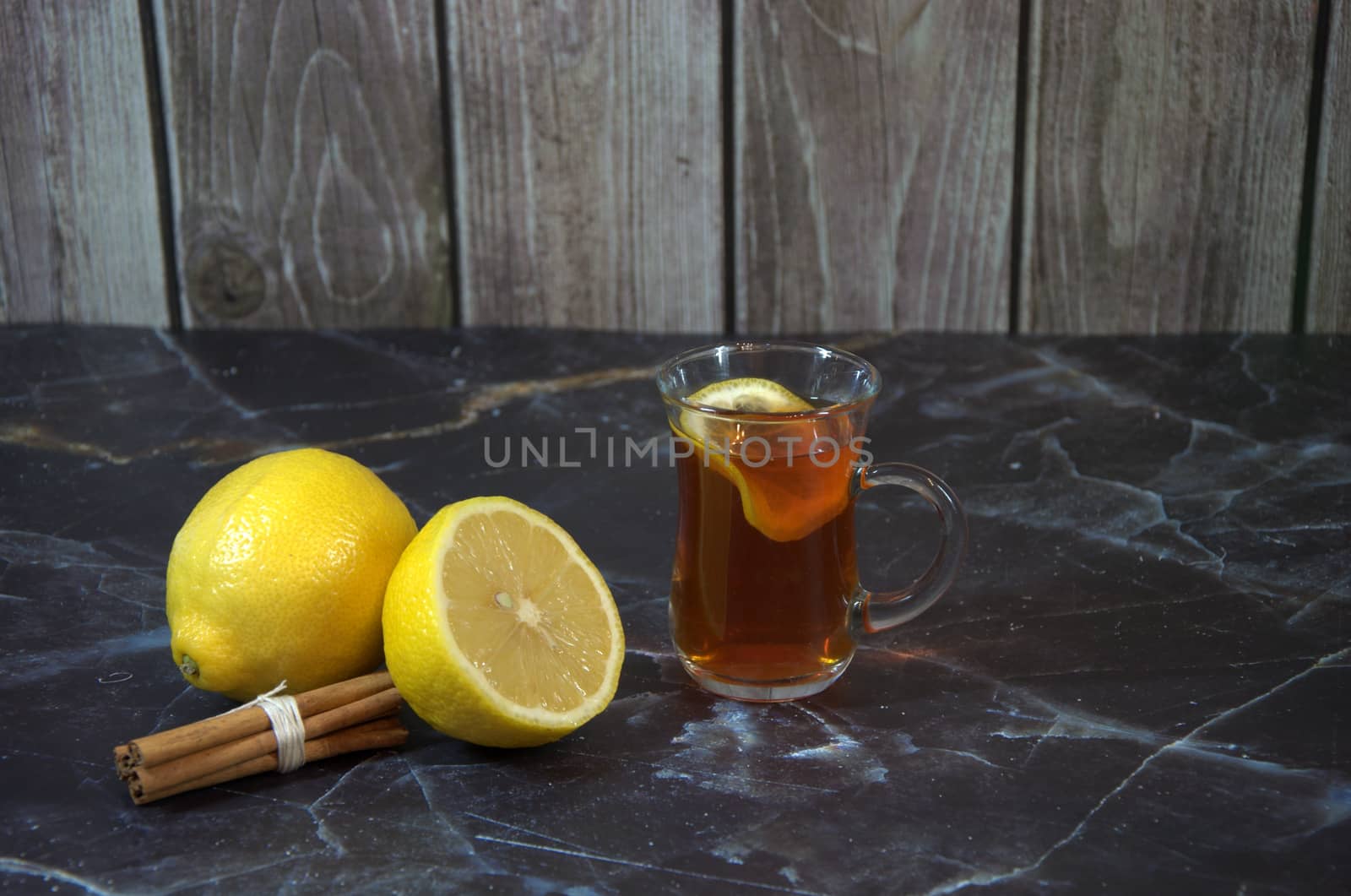 A glass cup of lemon tea with cinnamon sticks and a whole and half ripe lemon.