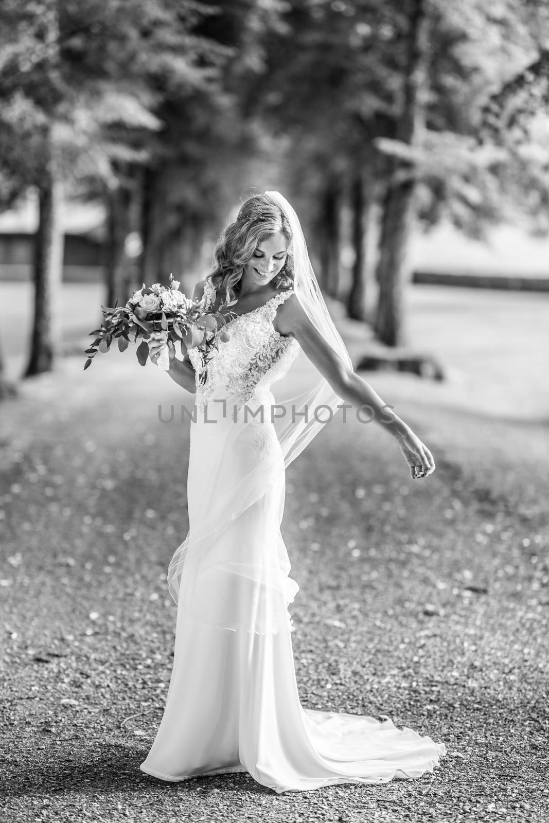 Full length portrait of beautiful sensual young blond bride in long white wedding dress and veil, holding bouquet outdoors in natural background. Black and white photo.