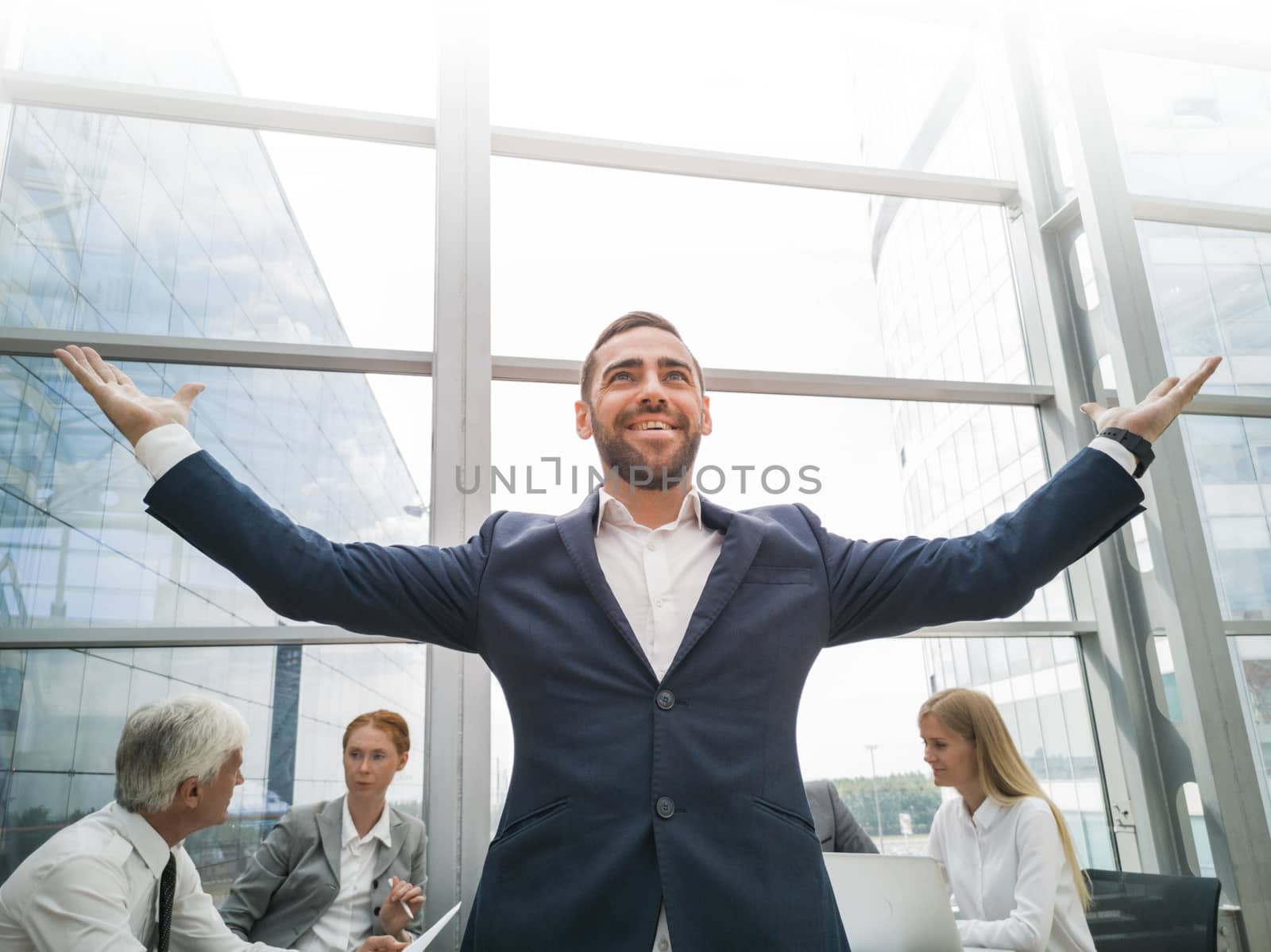 Young joyful businessman standing with arms raised smiling , team of his colleagues sitting at the table in the background