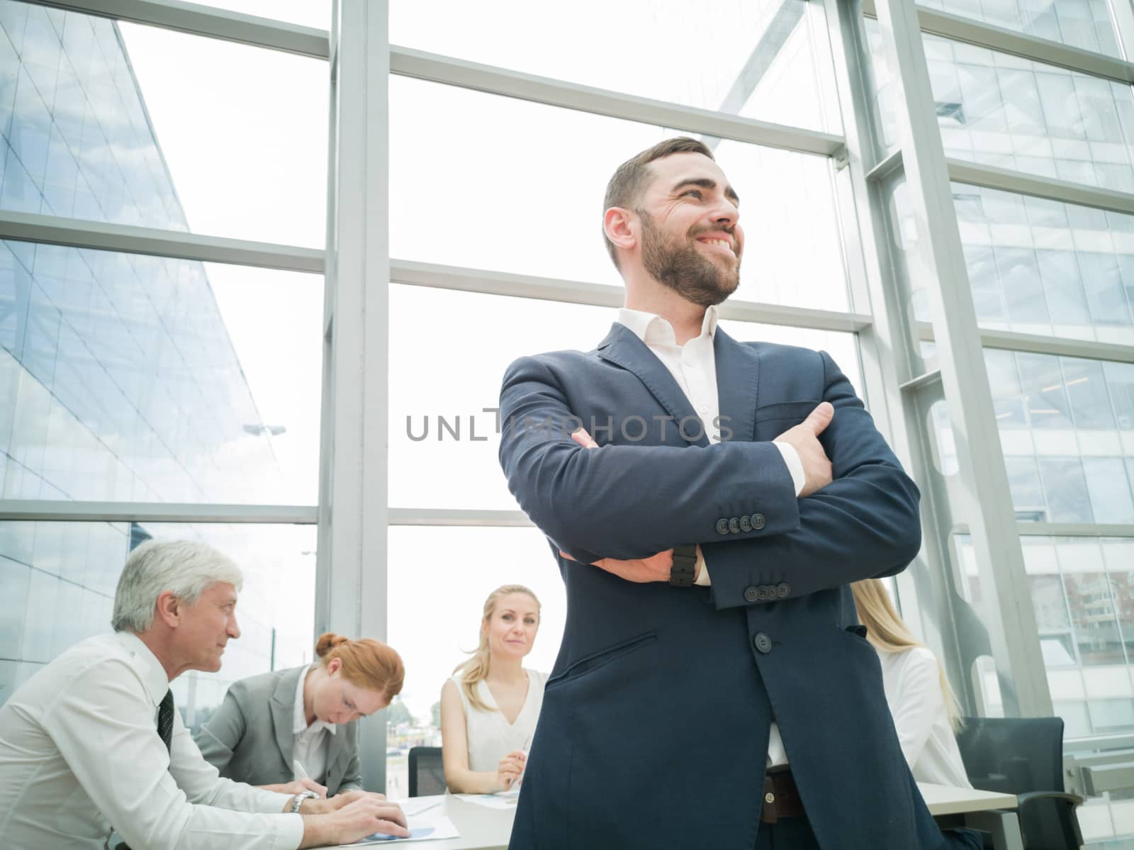 Confident young businessman standing with folded arms smiling , team of his colleagues sitting at the table in the background
