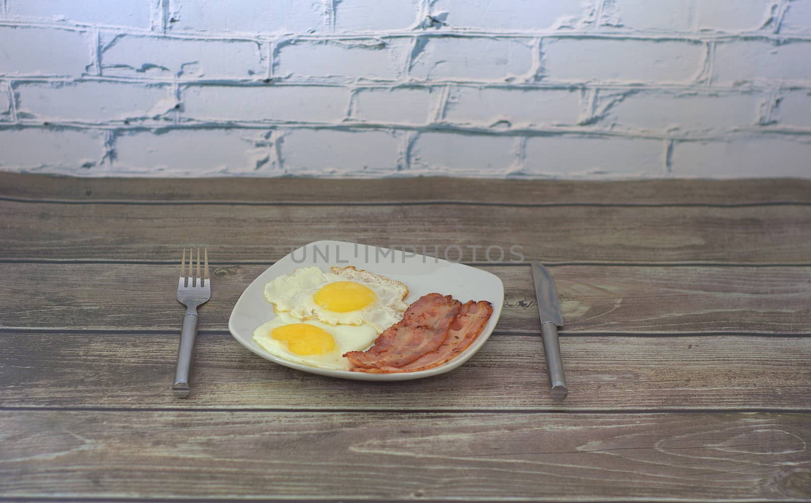 Breakfast, on a plate are two fried eggs and two slices of bacon in the form of an emoticon, next to a fork and knife.