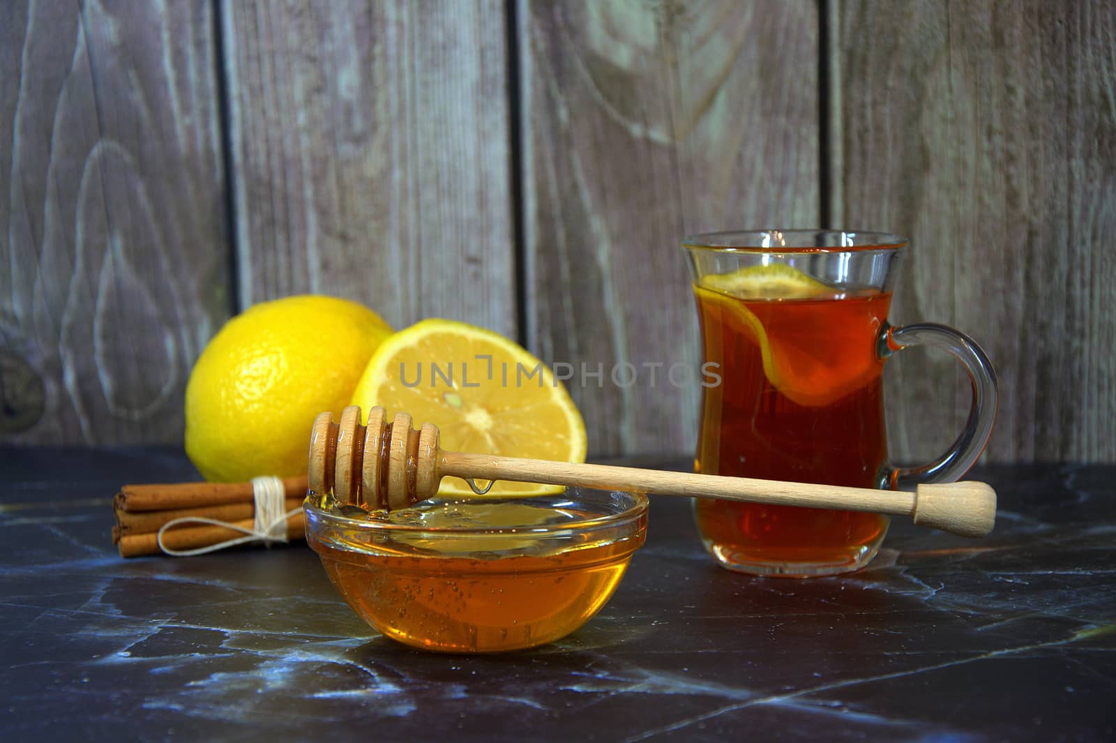 A glass of tea with lemon, a bowl of honey, two lemons and cinnamon sticks in the background.