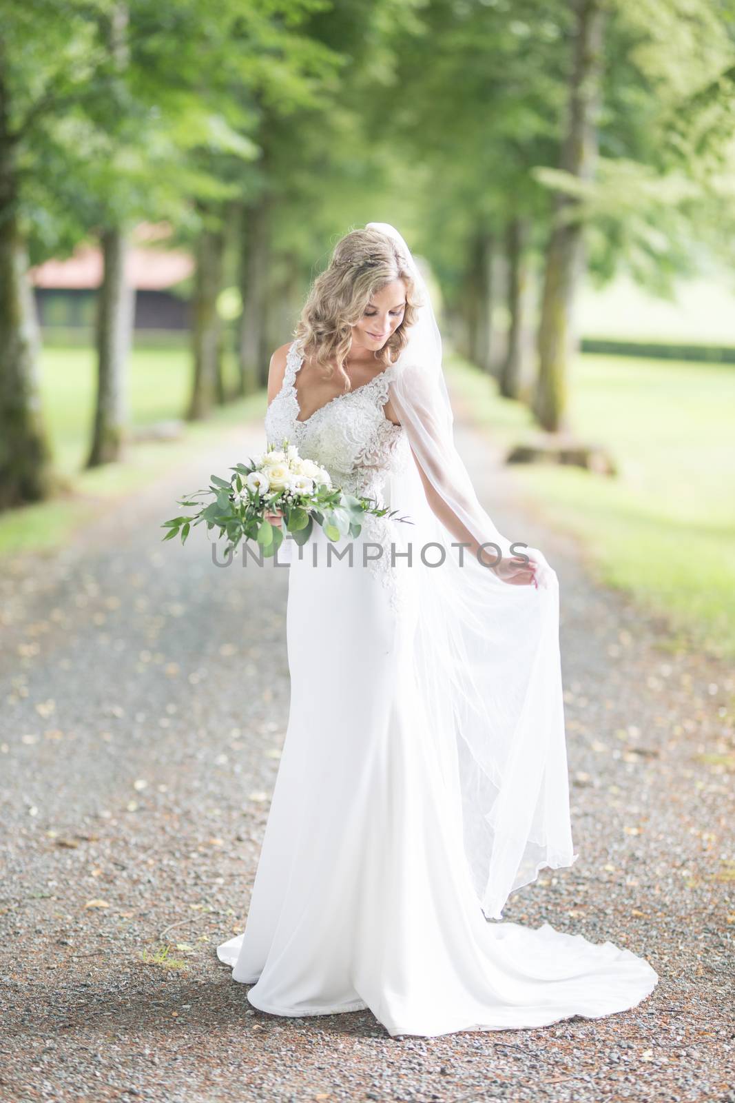 Full length portrait of beautiful sensual young blond bride in long white wedding dress and veil, holding bouquet outdoors in natural background.
