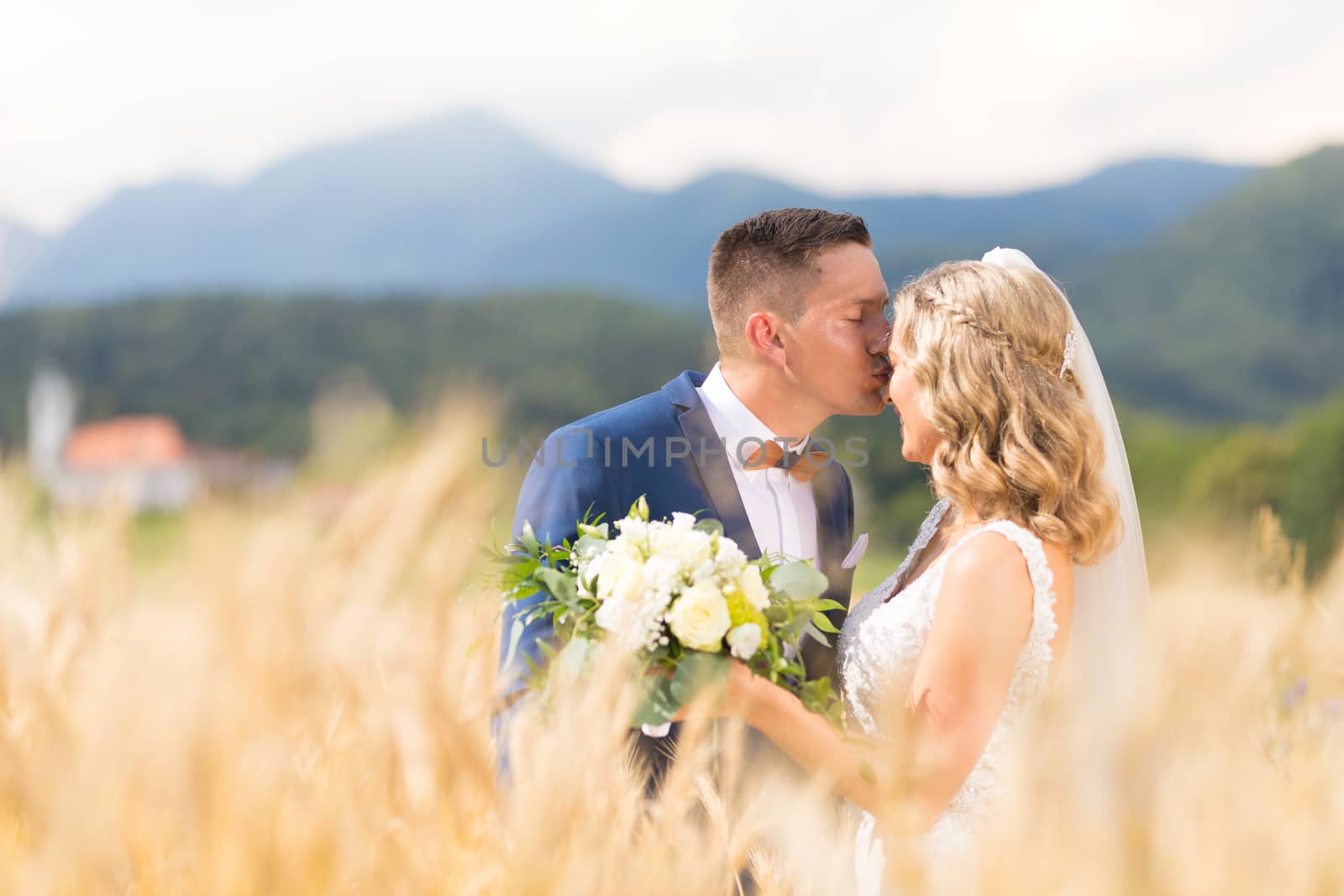 Groom hugging bride tenderly and kisses her on forehead in wheat field somewhere in Slovenian countryside. Caucasian happy romantic young couple celebrating their marriage.