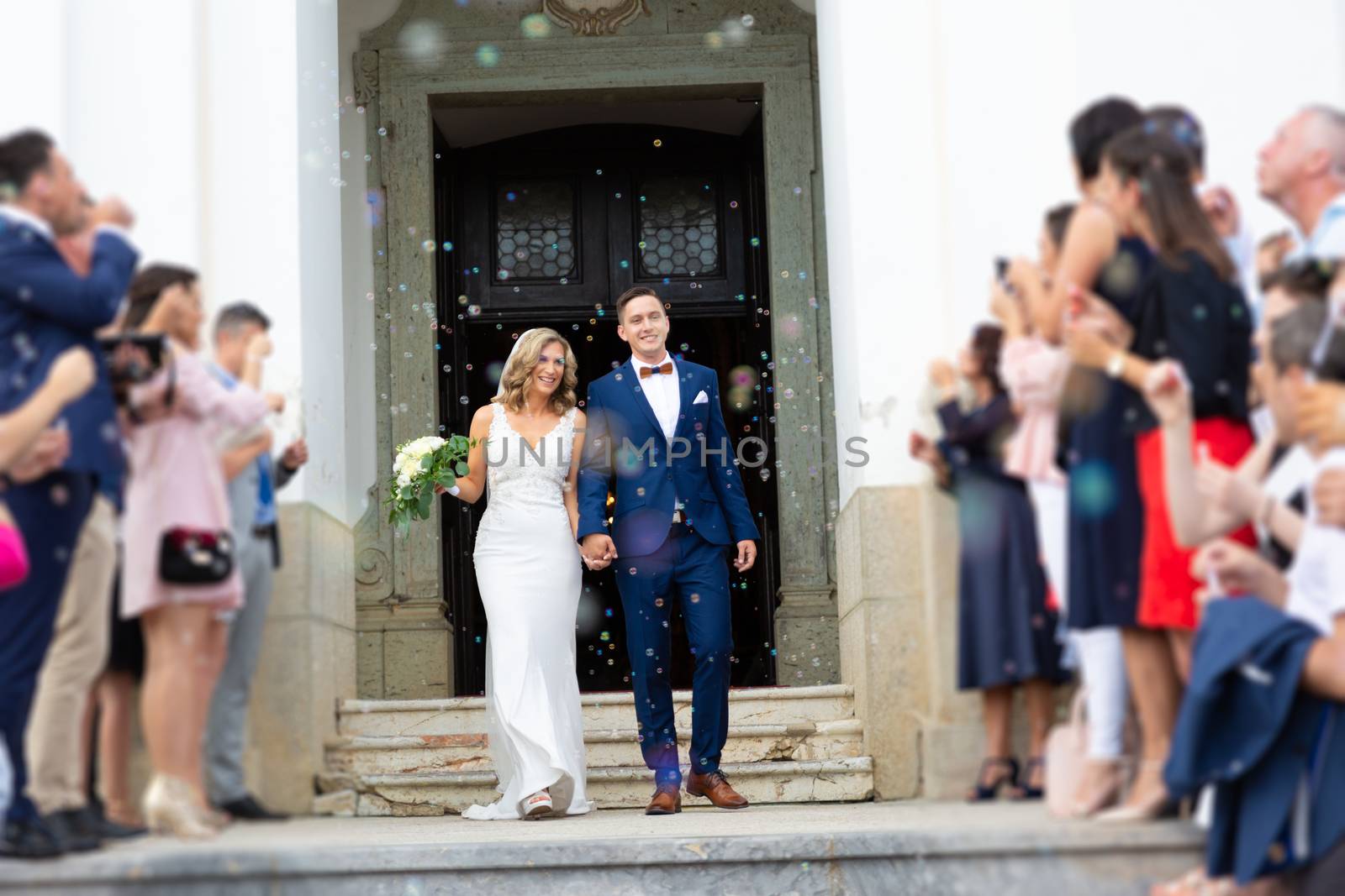 Newlyweds exiting the church after the wedding ceremony, family and friends celebrating their love with the shower of soap bubbles, custom undermining traditional rice bath.